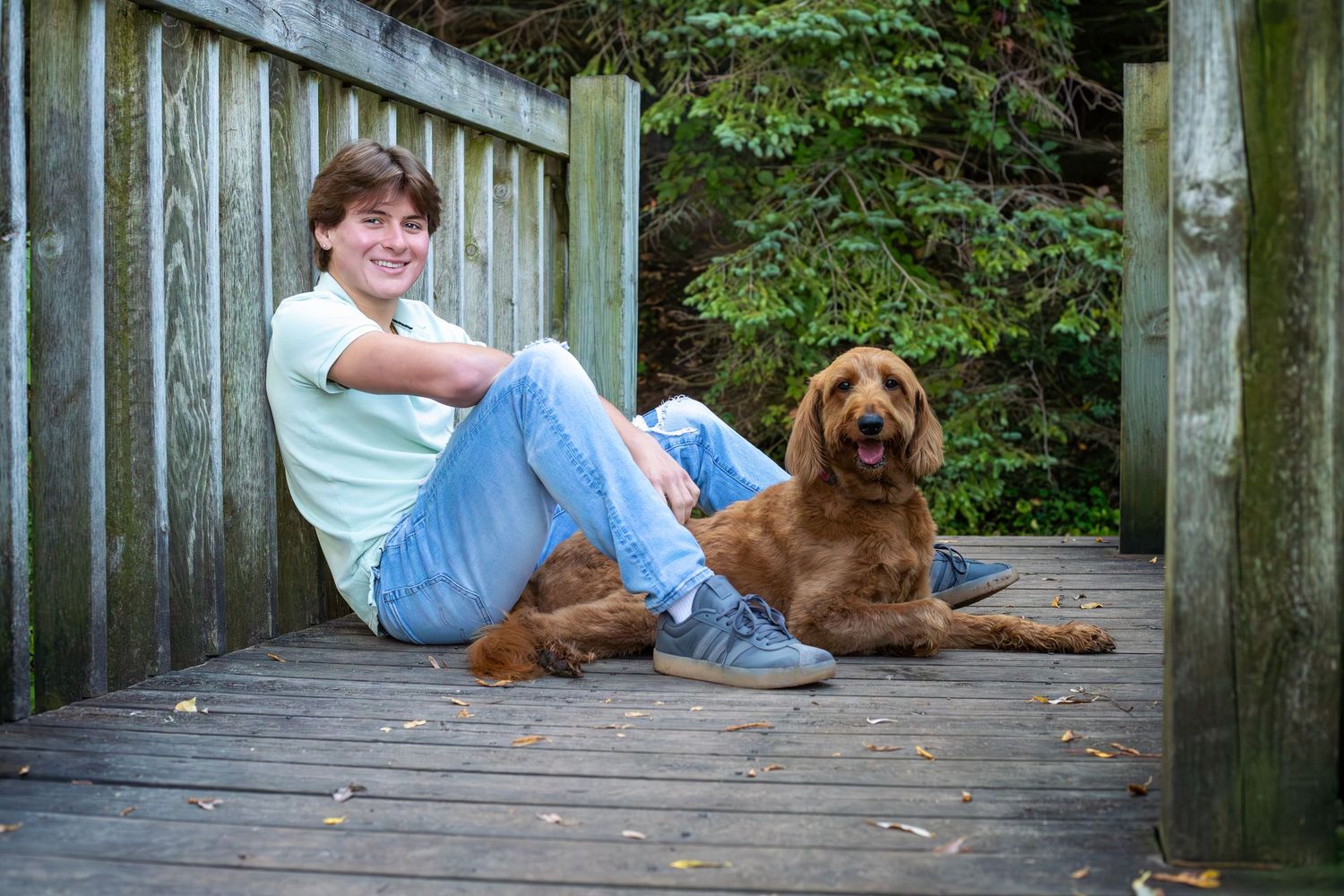 A brown dog and someone relaxing on a wooden deck surrounded by greenery.