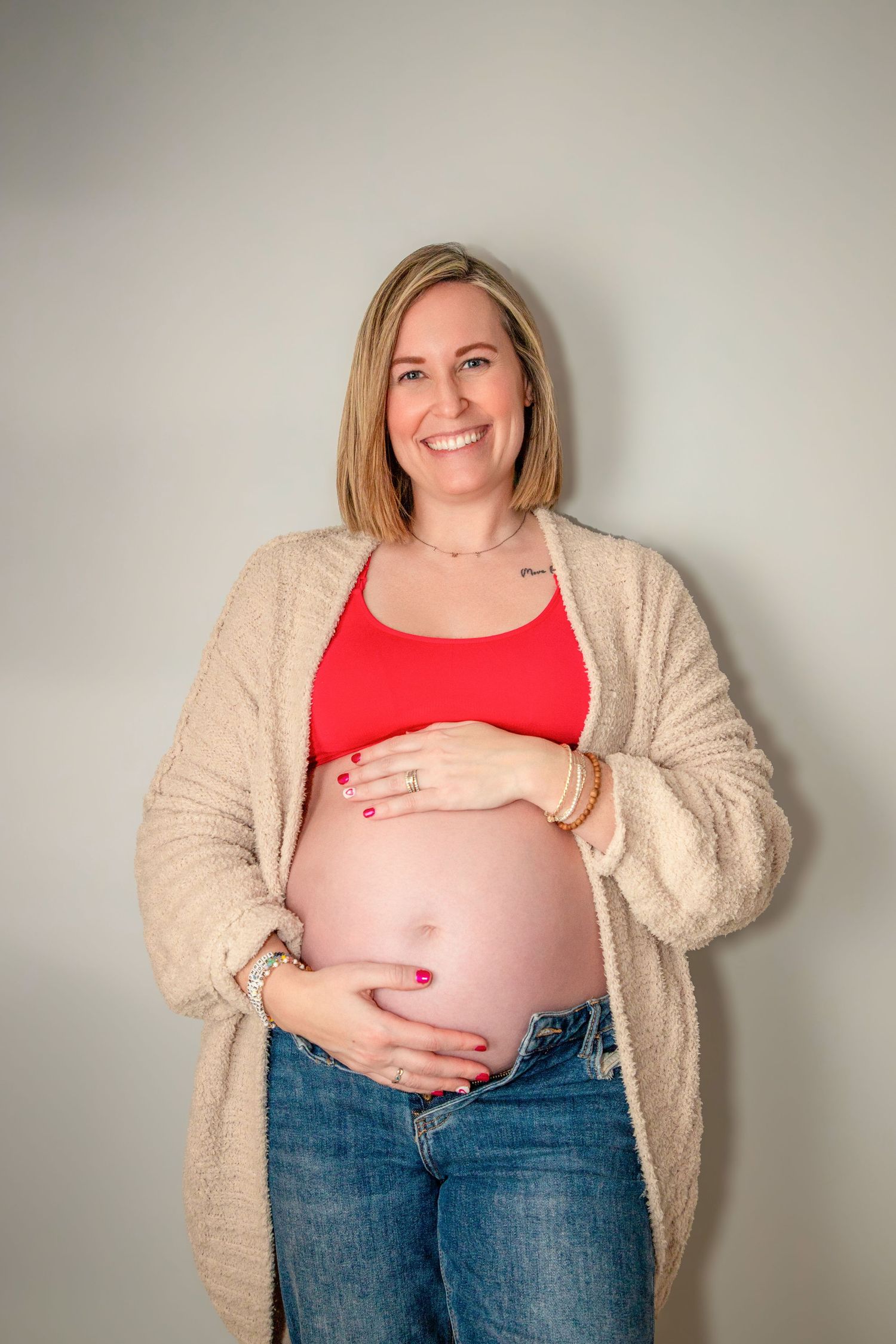 An expectant mother in a beige cardigan and red top cradles her baby bump against a neutral wall.