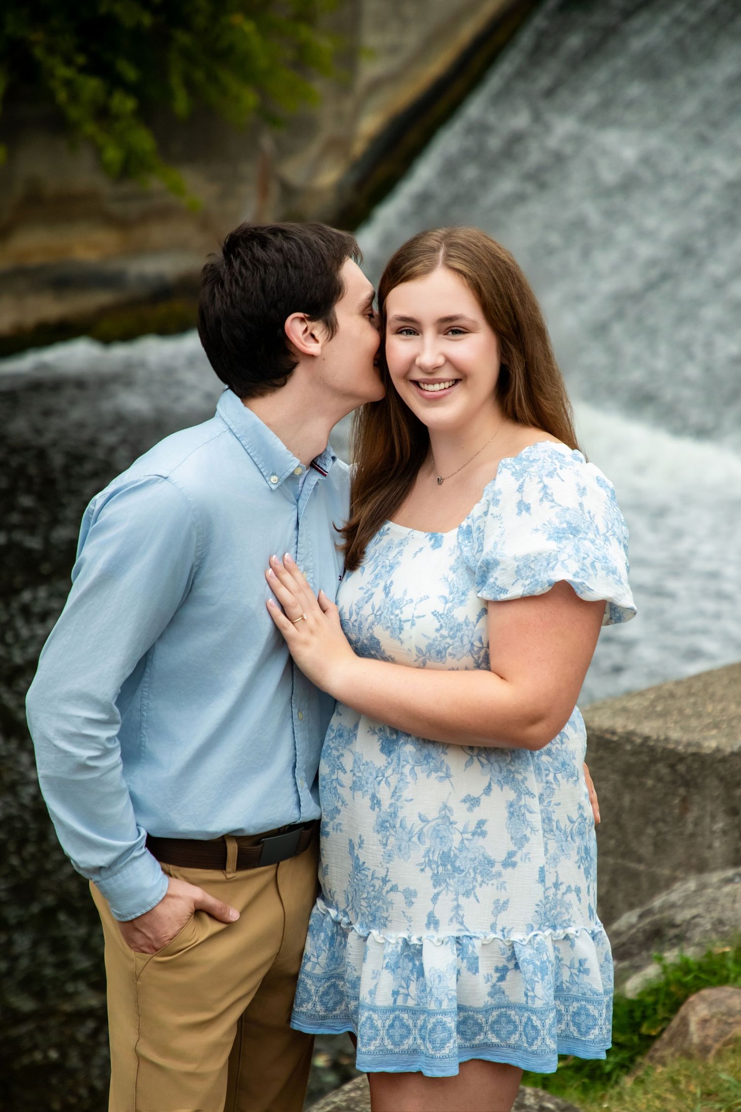 Romantic engagement photo of couple by waterfall with woman in blue floral dress.