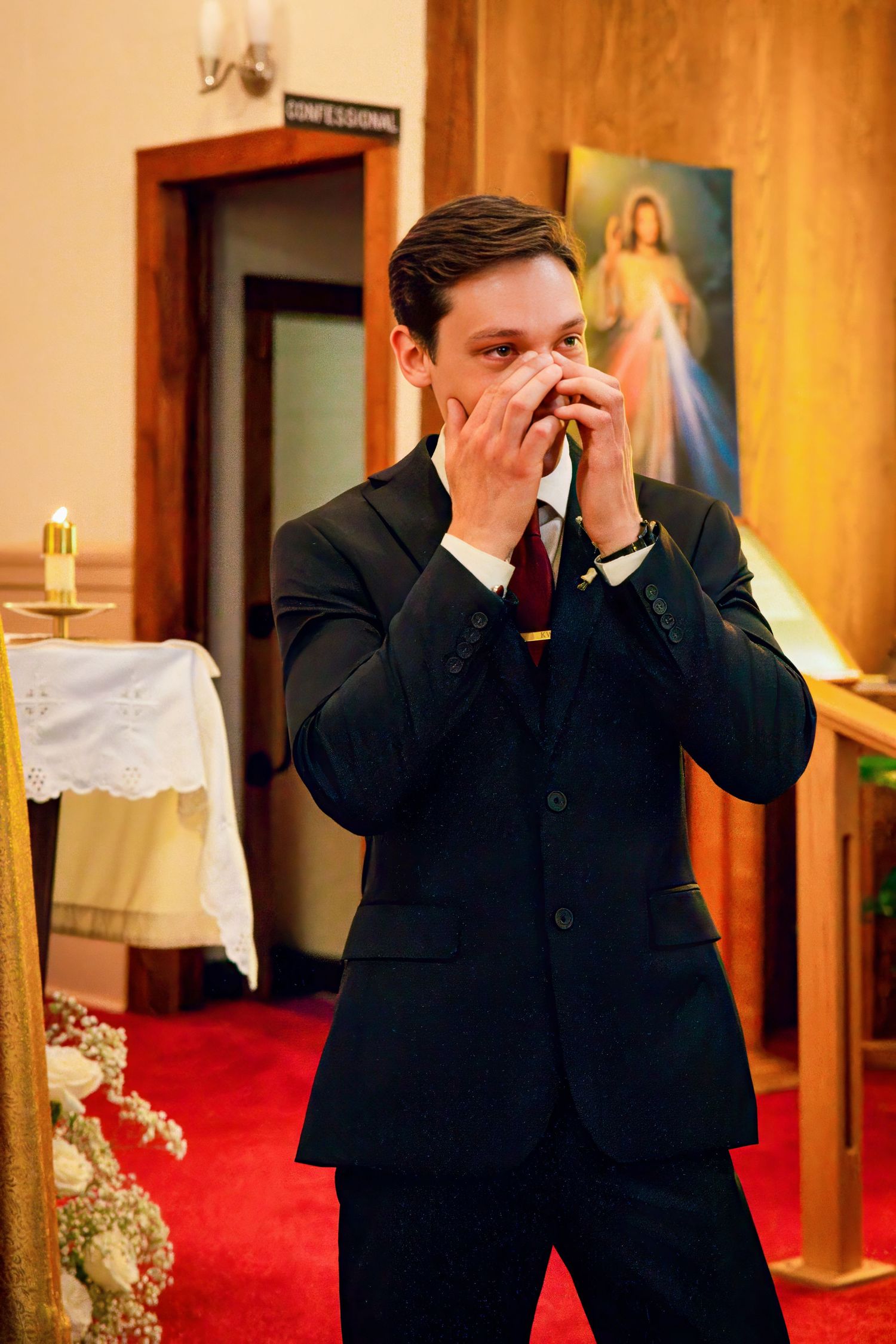 Man in dark suit shows emotion while covering his mouth in a church setting with a Divine Mercy painting visible.