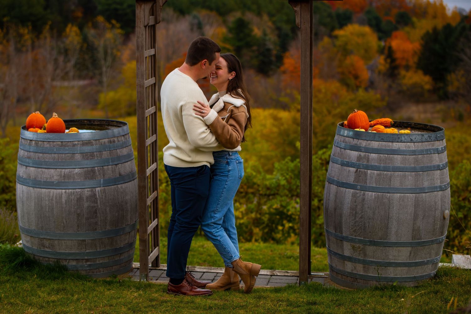 Romantic autumn engagement photo session with wine barrels and fall foliage in rustic outdoor setting.