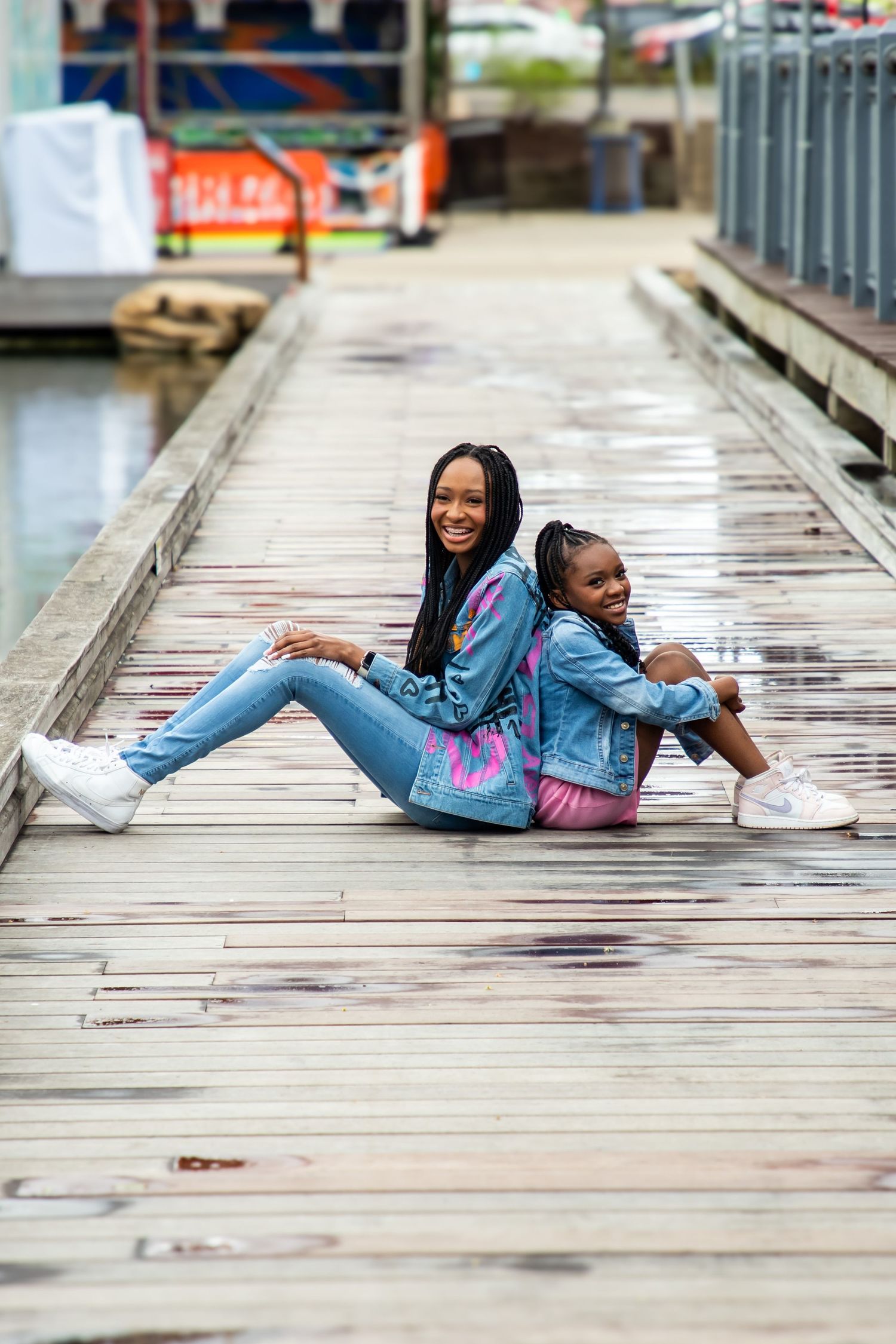 Two people in matching denim outfits sit back-to-back on wooden dock.