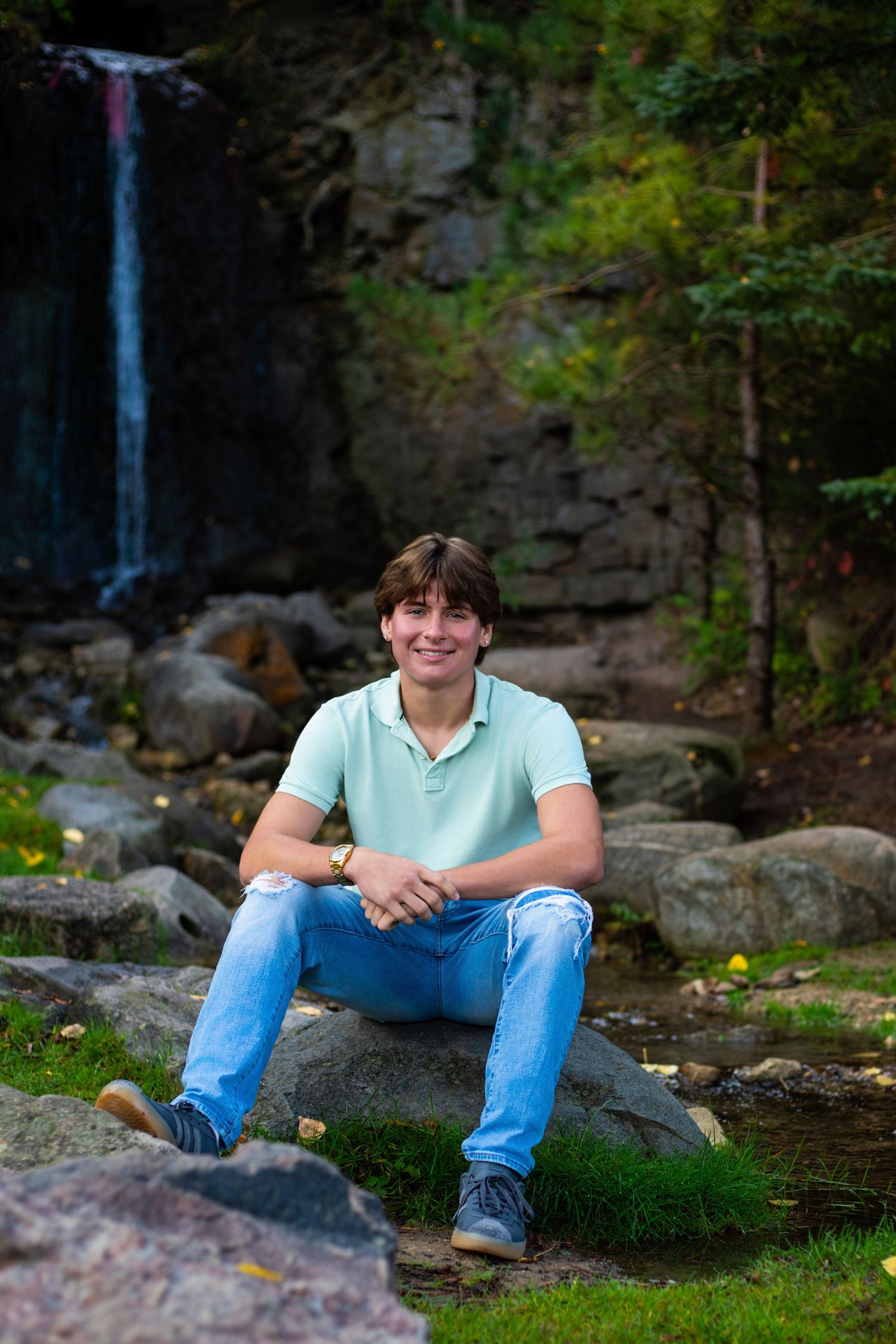 Person in polo shirt and jeans sits on rock near scenic waterfall.