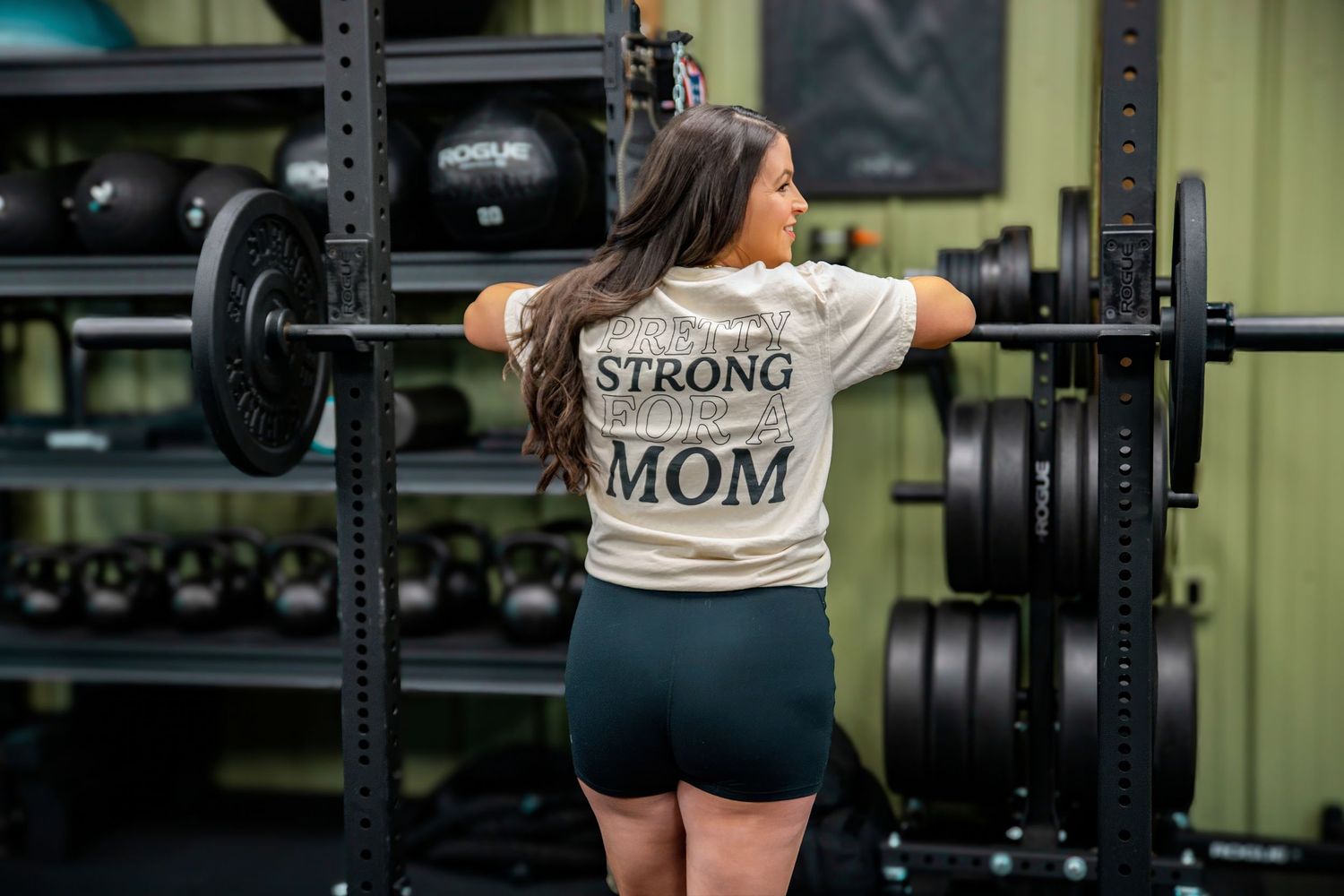 Athletic woman in gym wearing 'Strong Mom' shirt loading weights on squat rack.