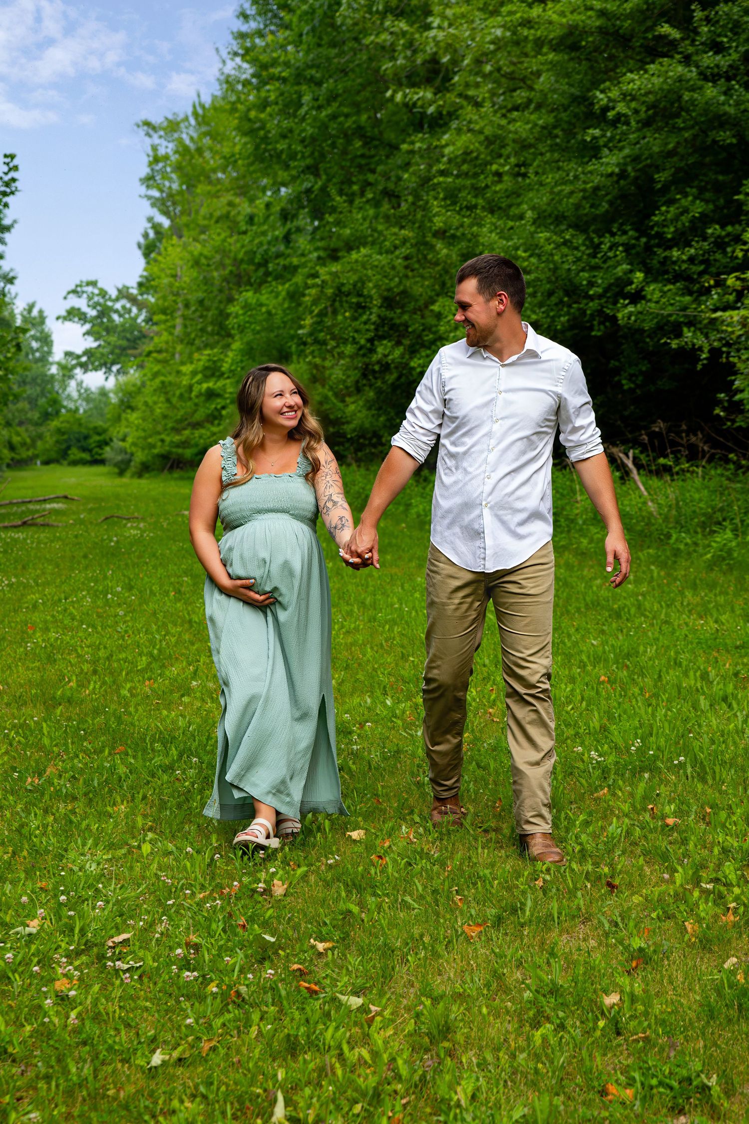 Couple walking hand in hand through green summer meadow during maternity photoshoot.