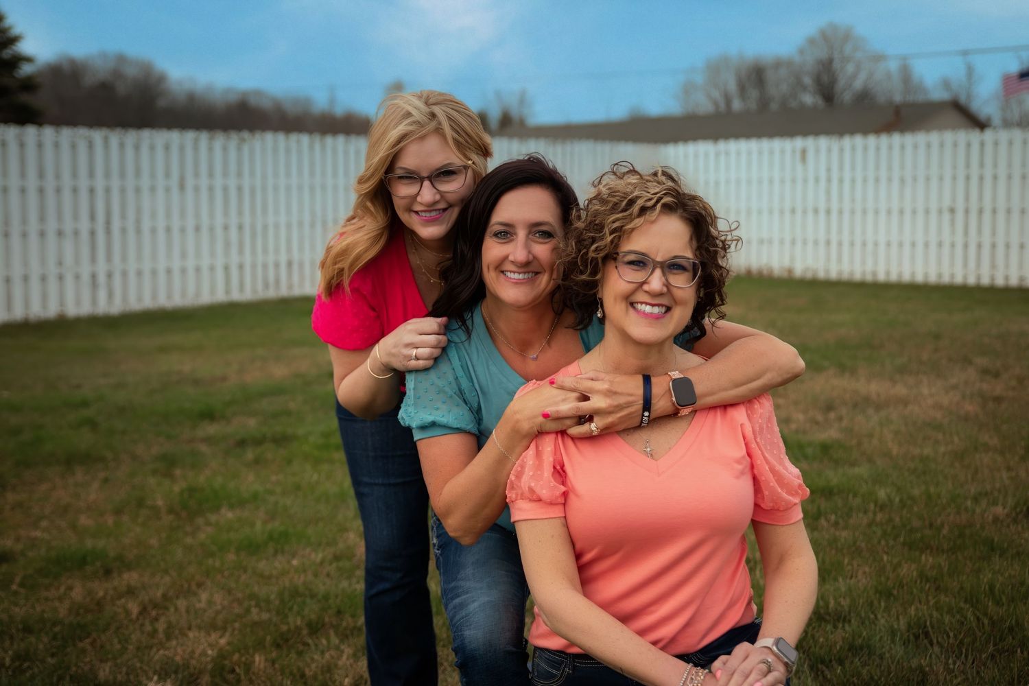 Three people posing together outdoors in casual attire against a white fence on a cloudy day.