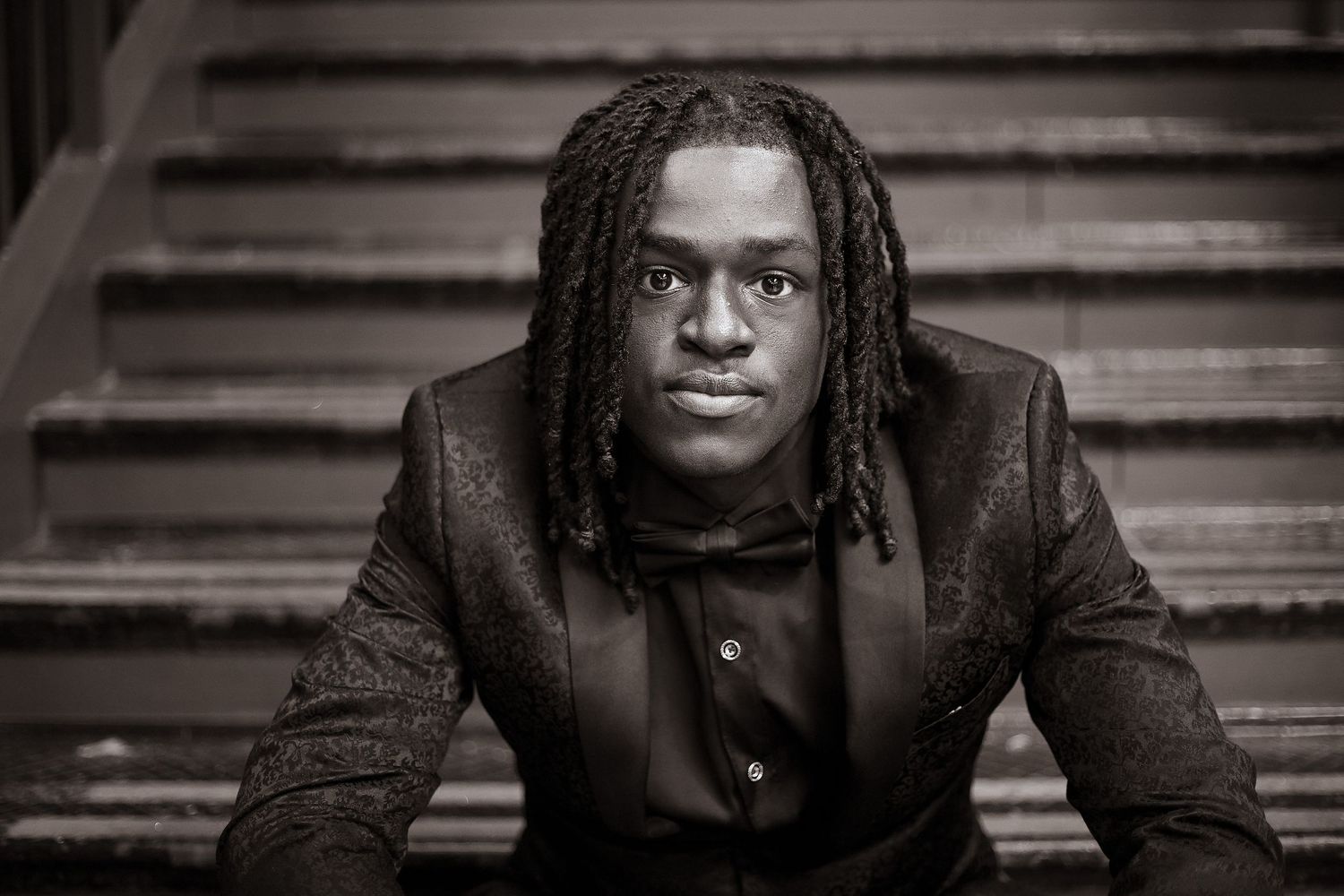 Dramatic black and white portrait of person in dark jacket with dreadlocks against wooden steps background.