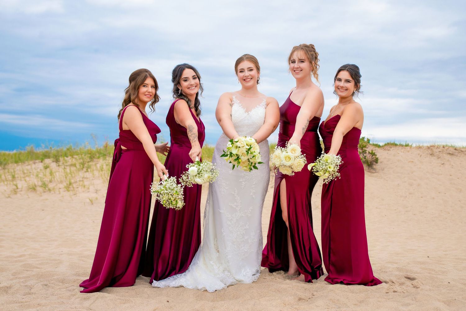 Wedding party on beach with bride in white gown surrounded by bridesmaids in burgundy dresses.