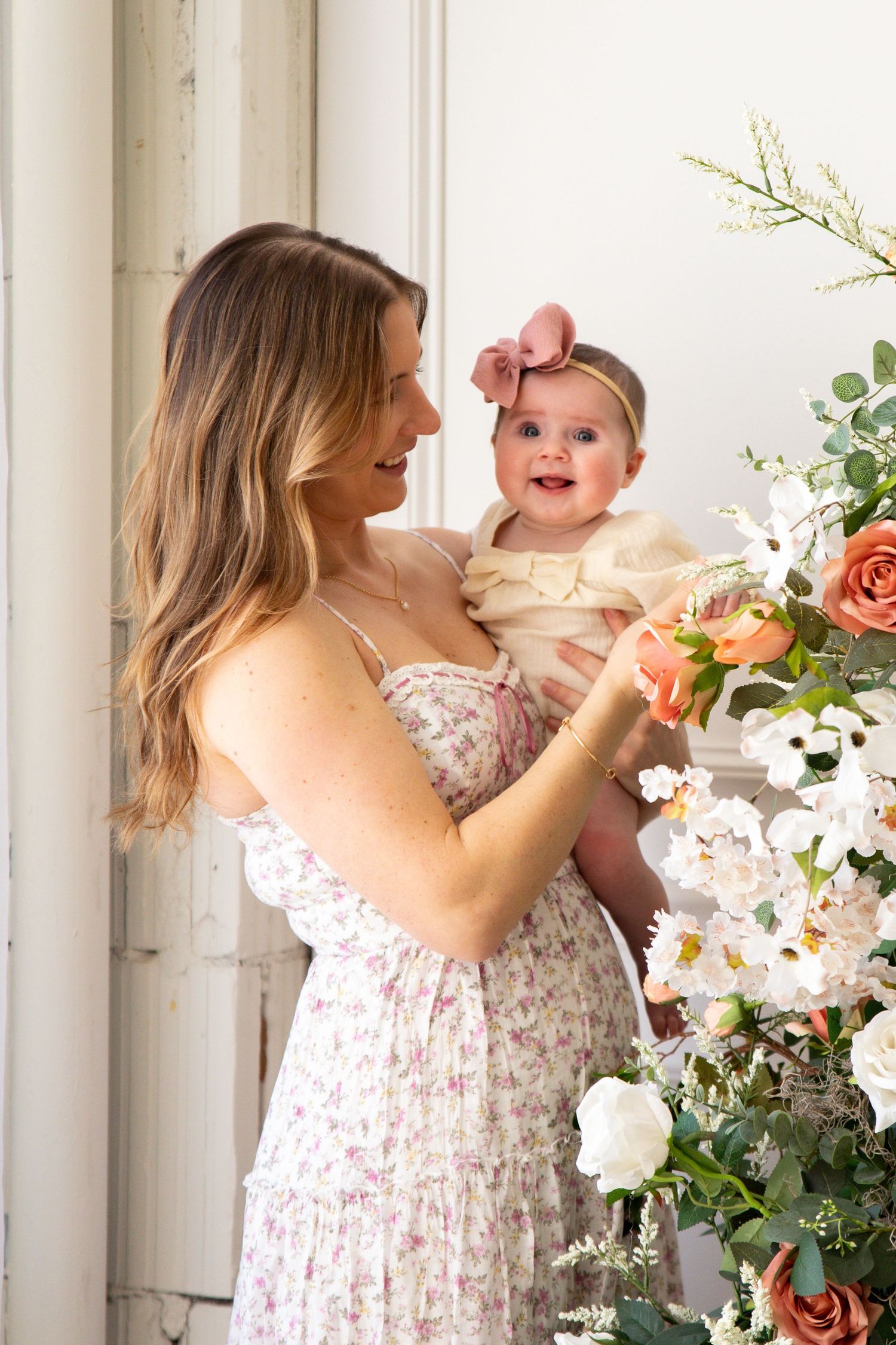 Mother and baby sharing tender moment by floral arrangement in bright white room.