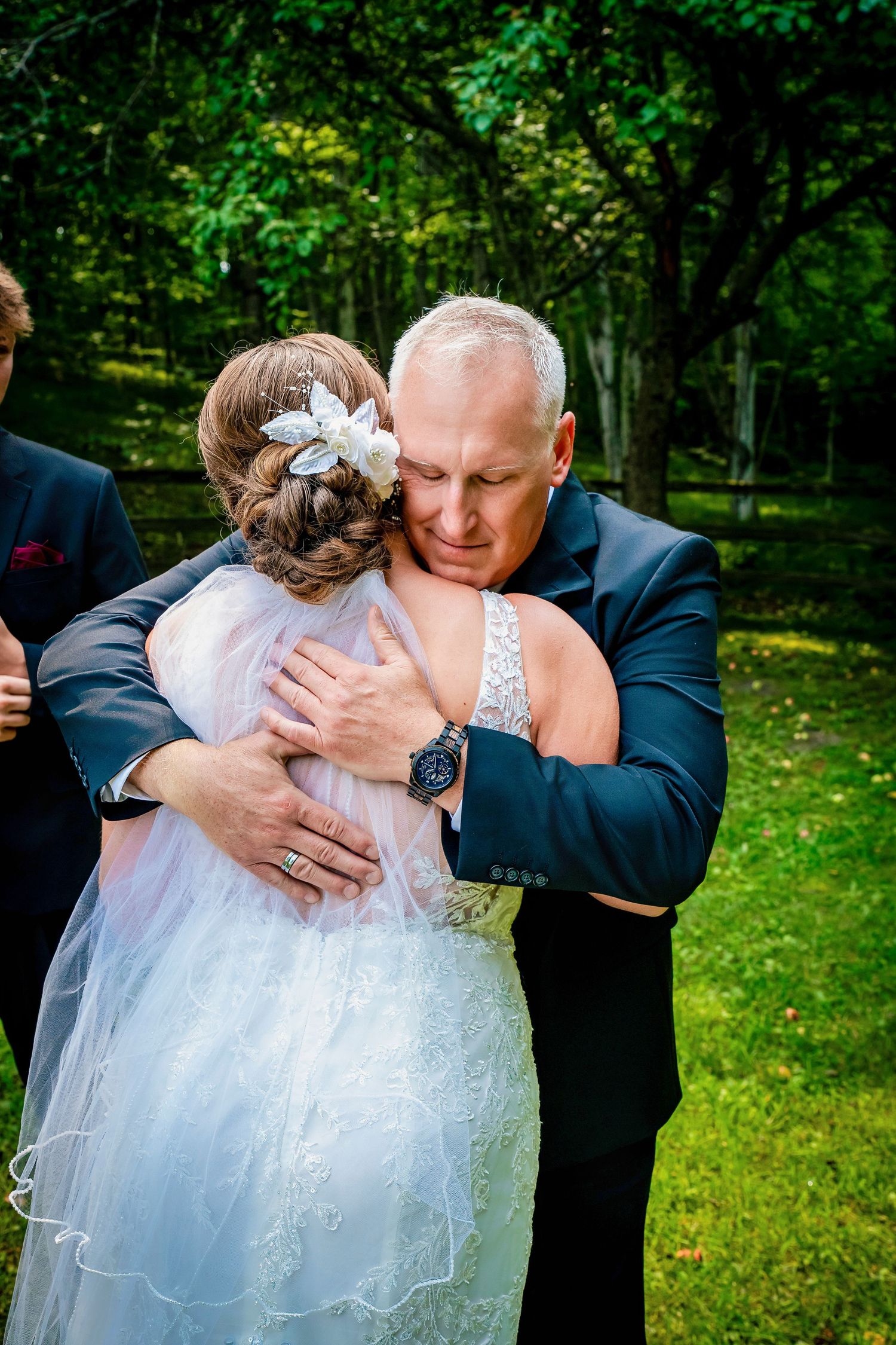A tender moment captured at an outdoor wedding with a couple embracing against a lush green forest backdrop.
