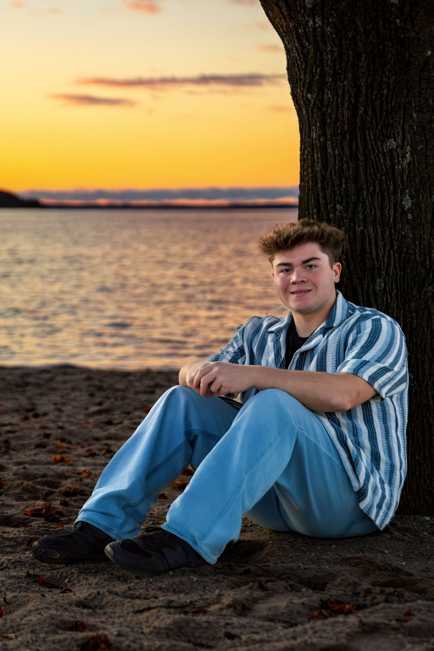 Casual portrait at sunset of person sitting against tree by waterfront.