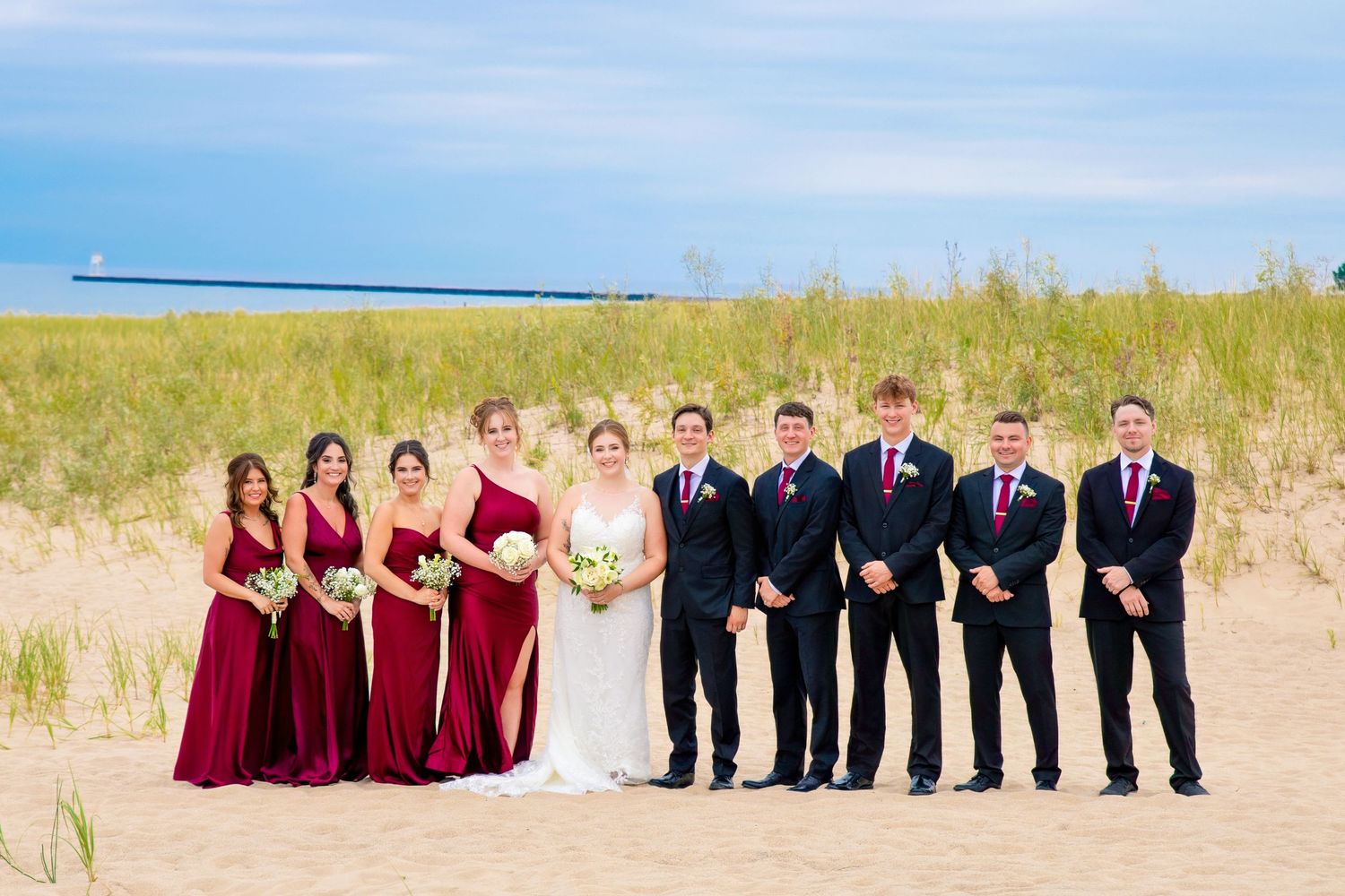 Wedding party in burgundy dresses and navy suits poses in a line formation on a sandy beach.