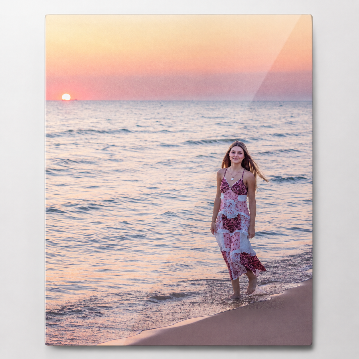 Young woman in floral dress walking on sandy beach at golden sunset with calm ocean waves in background.
