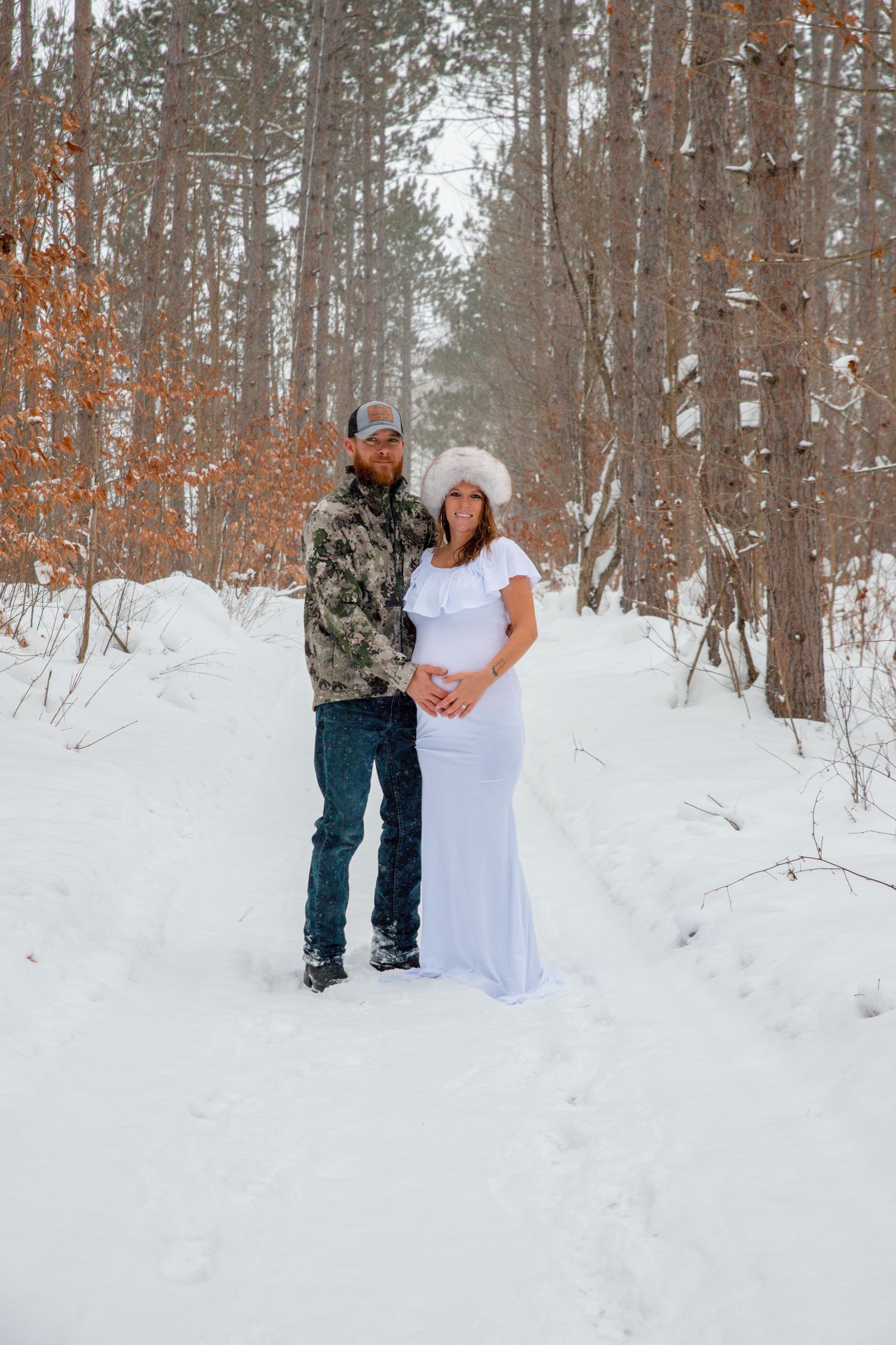 Snowy winter maternity photoshoot in a forest with couple in white dress and camo.