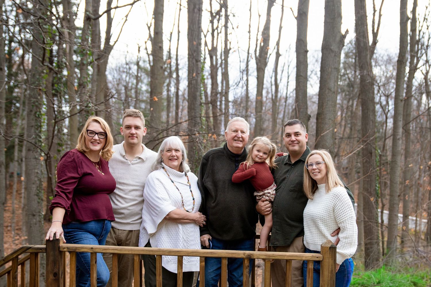 Group portrait of six people standing together on a wooden deck in a wooded setting during autumn.