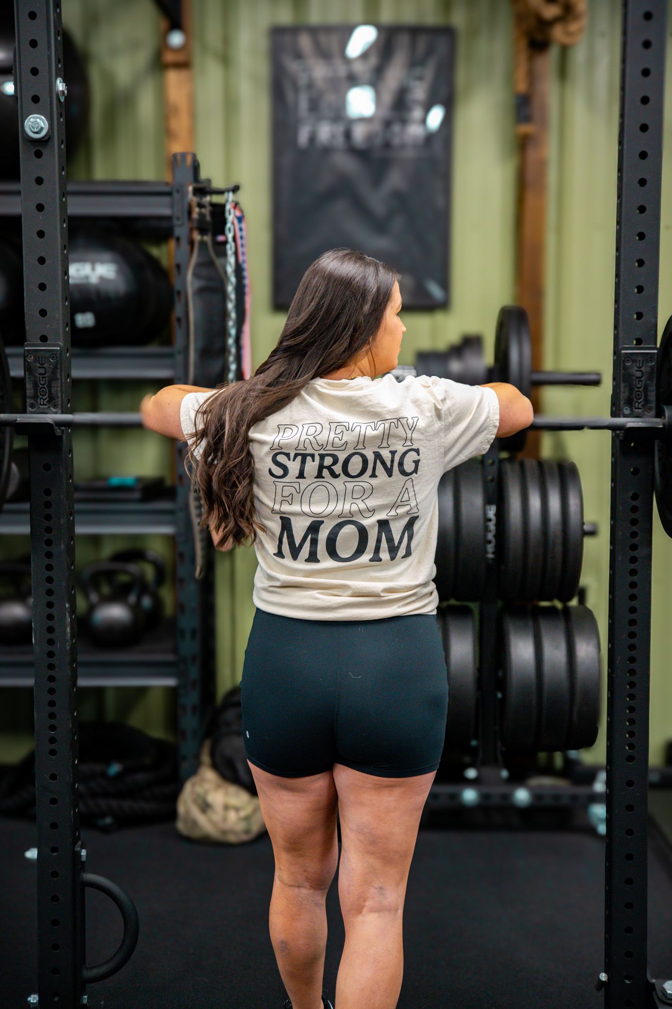Athletic woman in gym wearing 'Strong Mom' t-shirt preparing for workout at squat rack.
