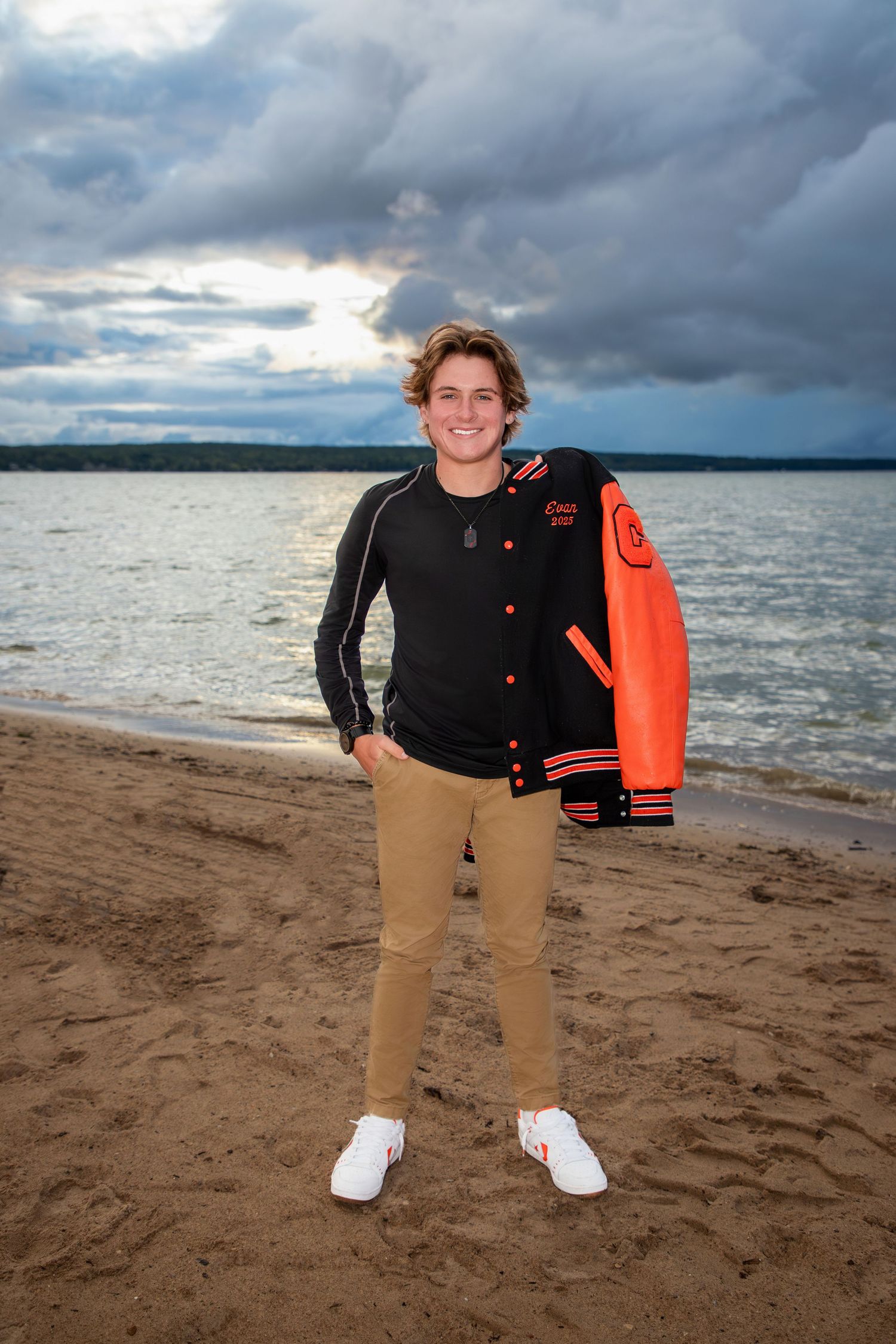 Person wearing black jacket and khakis poses on beach under stormy skies.