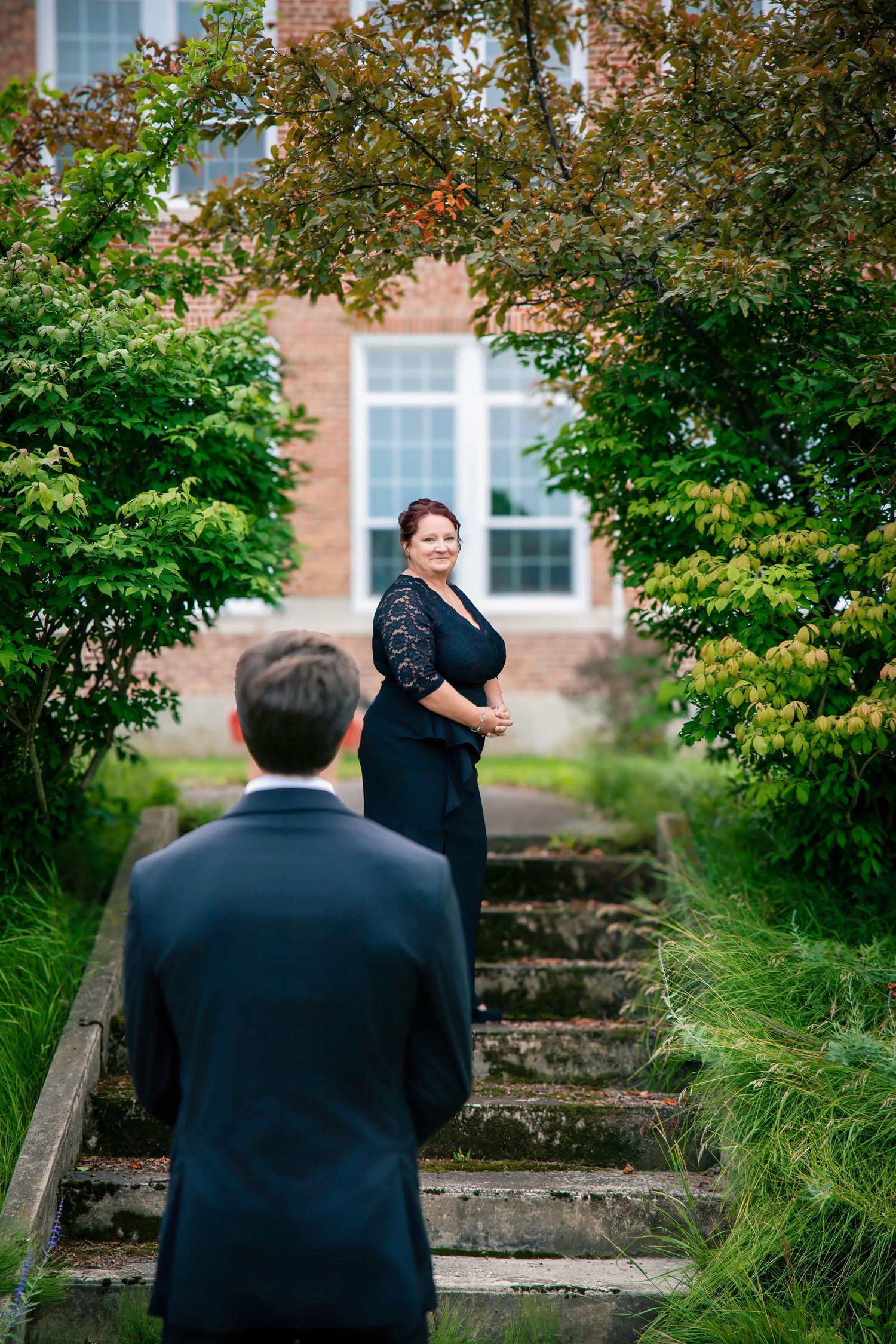 A couple in formal attire pose on old stone garden steps with green foliage and a brick building in the background.