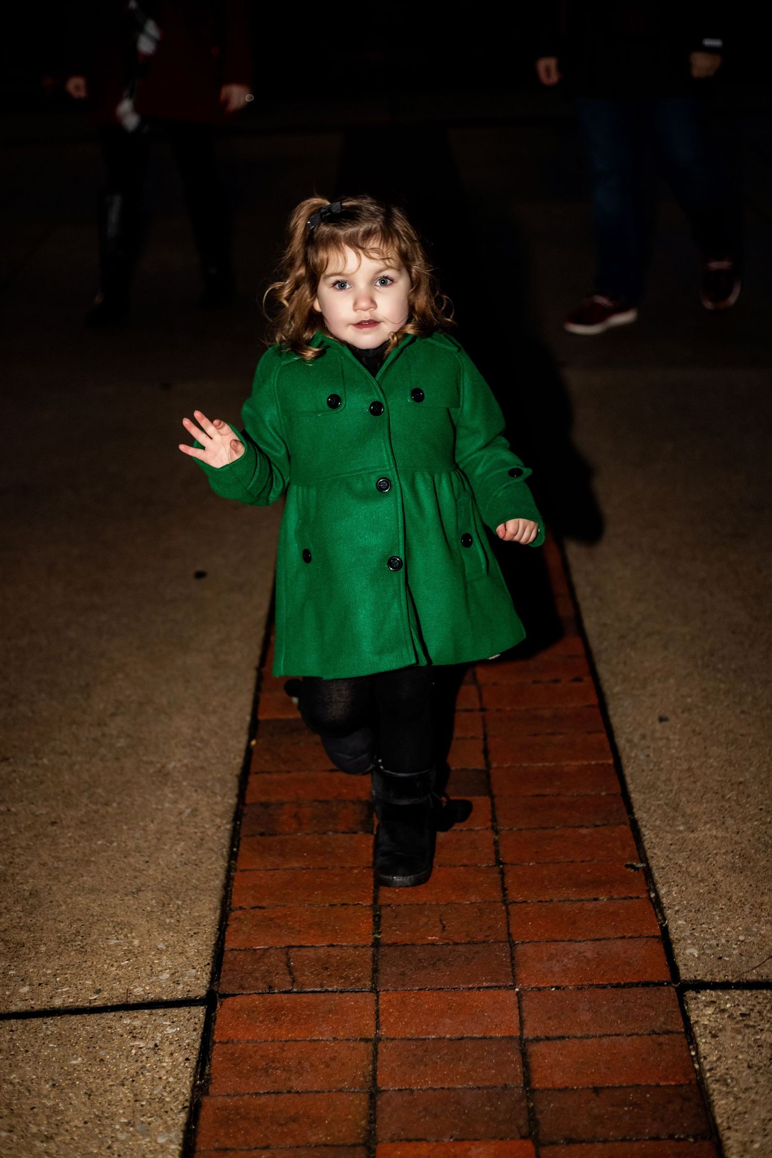 Small child in emerald green coat walking on brick pavement at night.