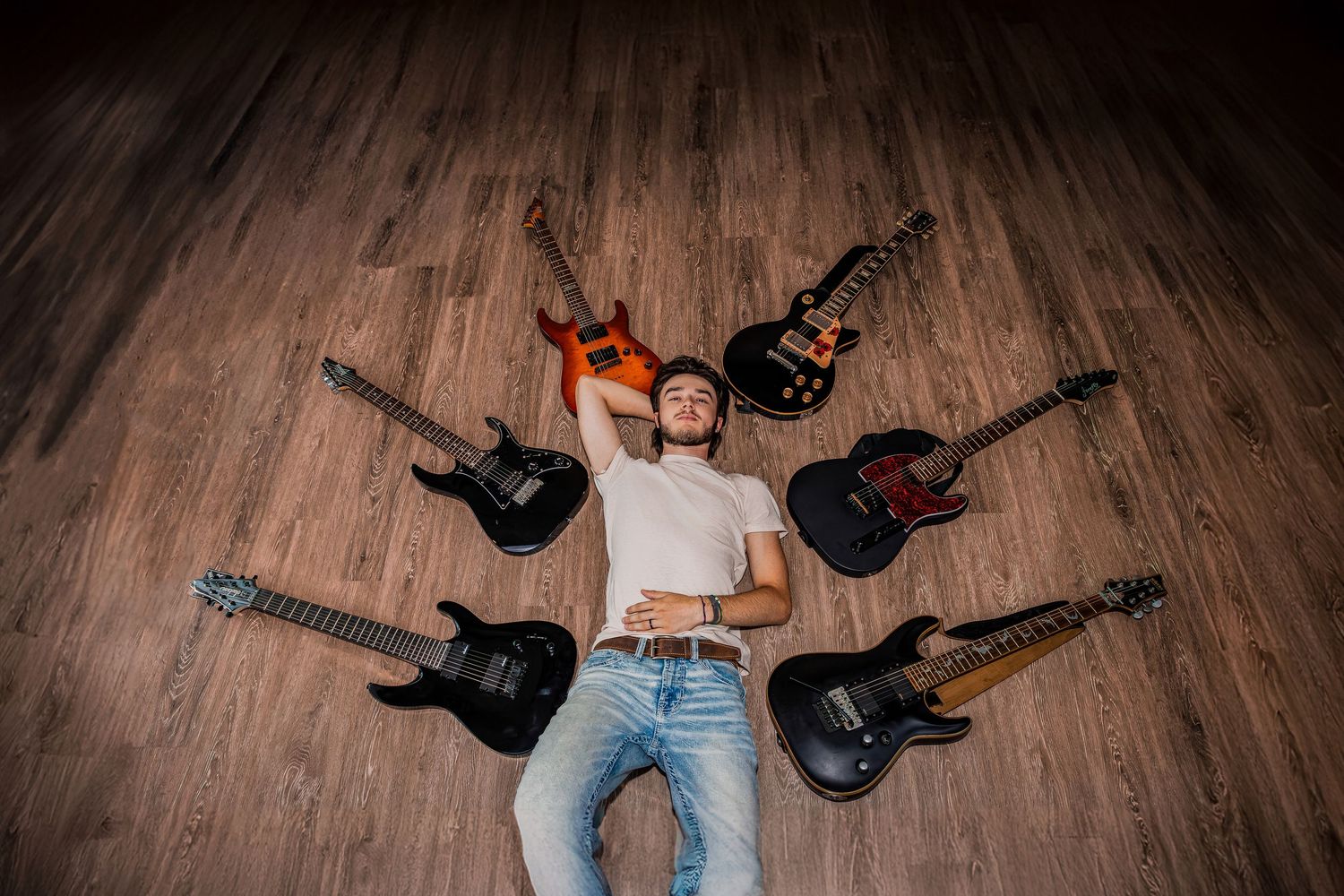 An artistic overhead shot of a person lying on wooden floor surrounded by guitars in a circle.