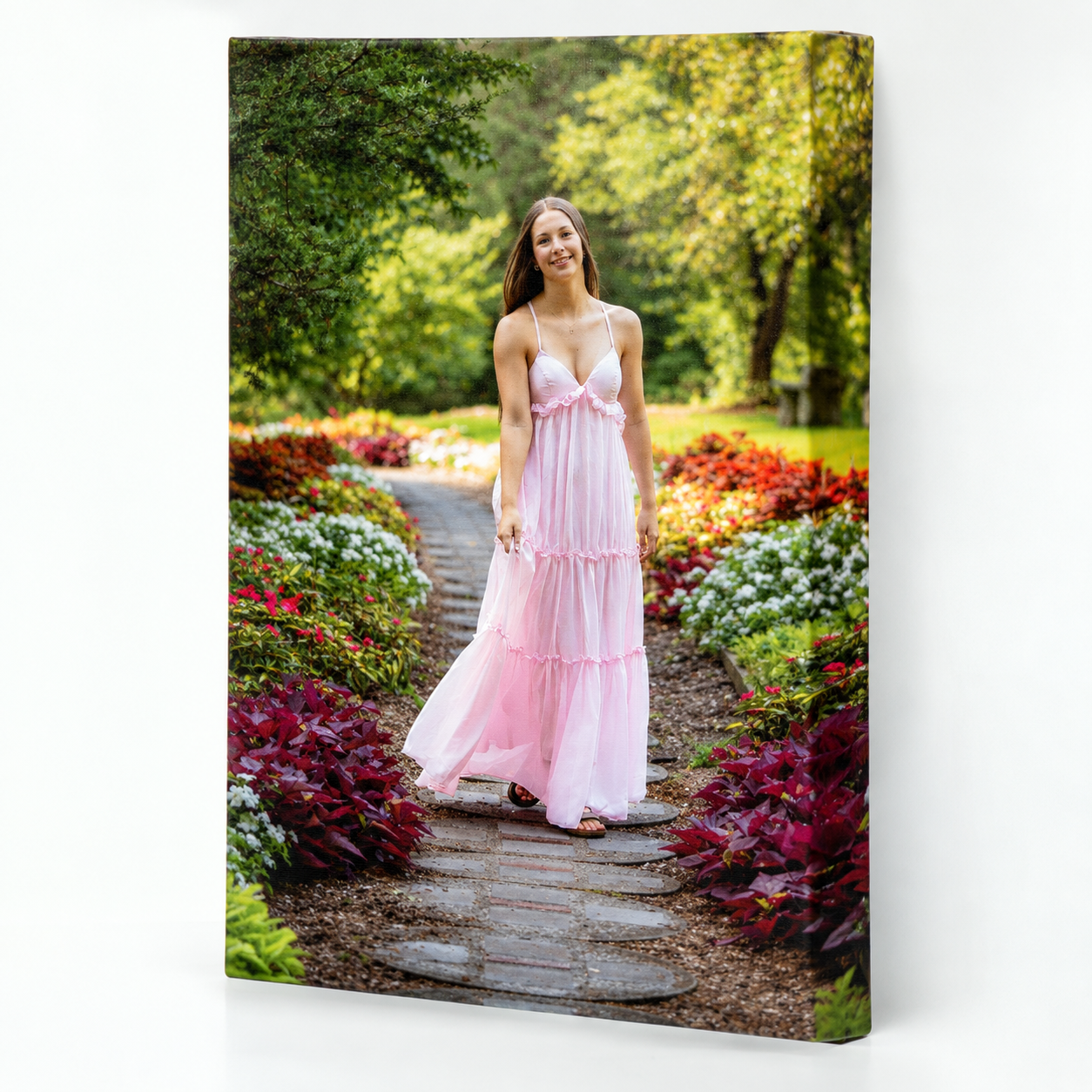 A young woman in a flowing pink dress stands on a stone path surrounded by colorful garden flowers.