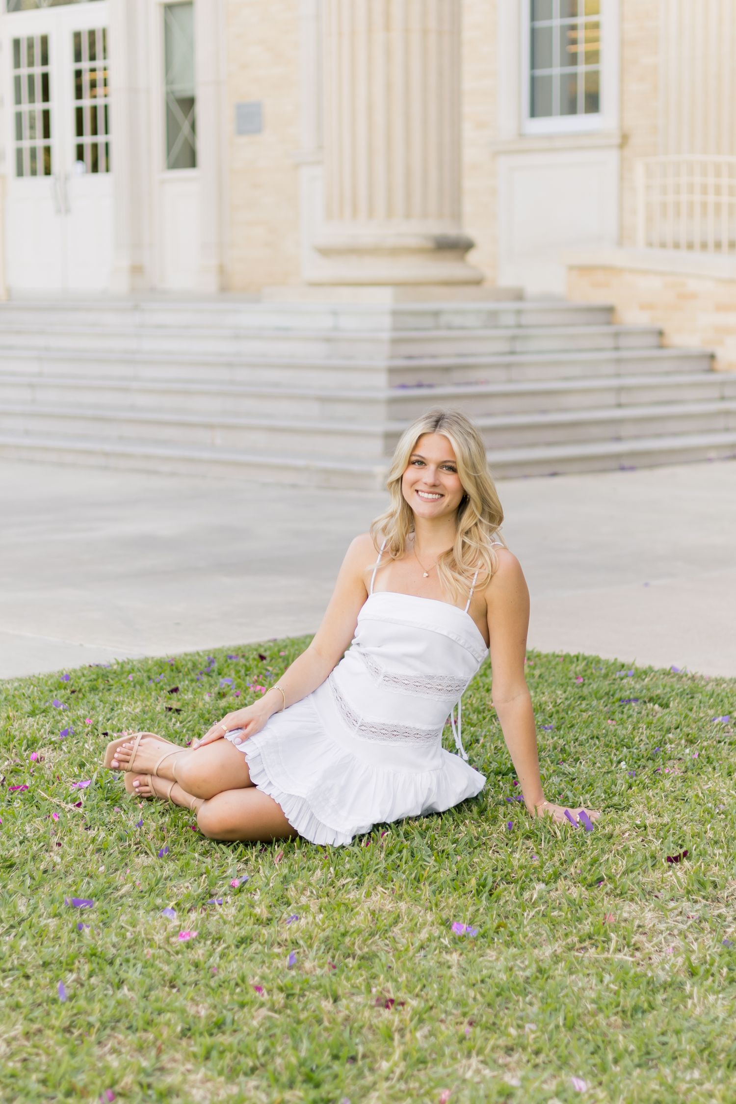 Smiling blonde woman in white mini dress sitting barefoot on green grass near a university building.