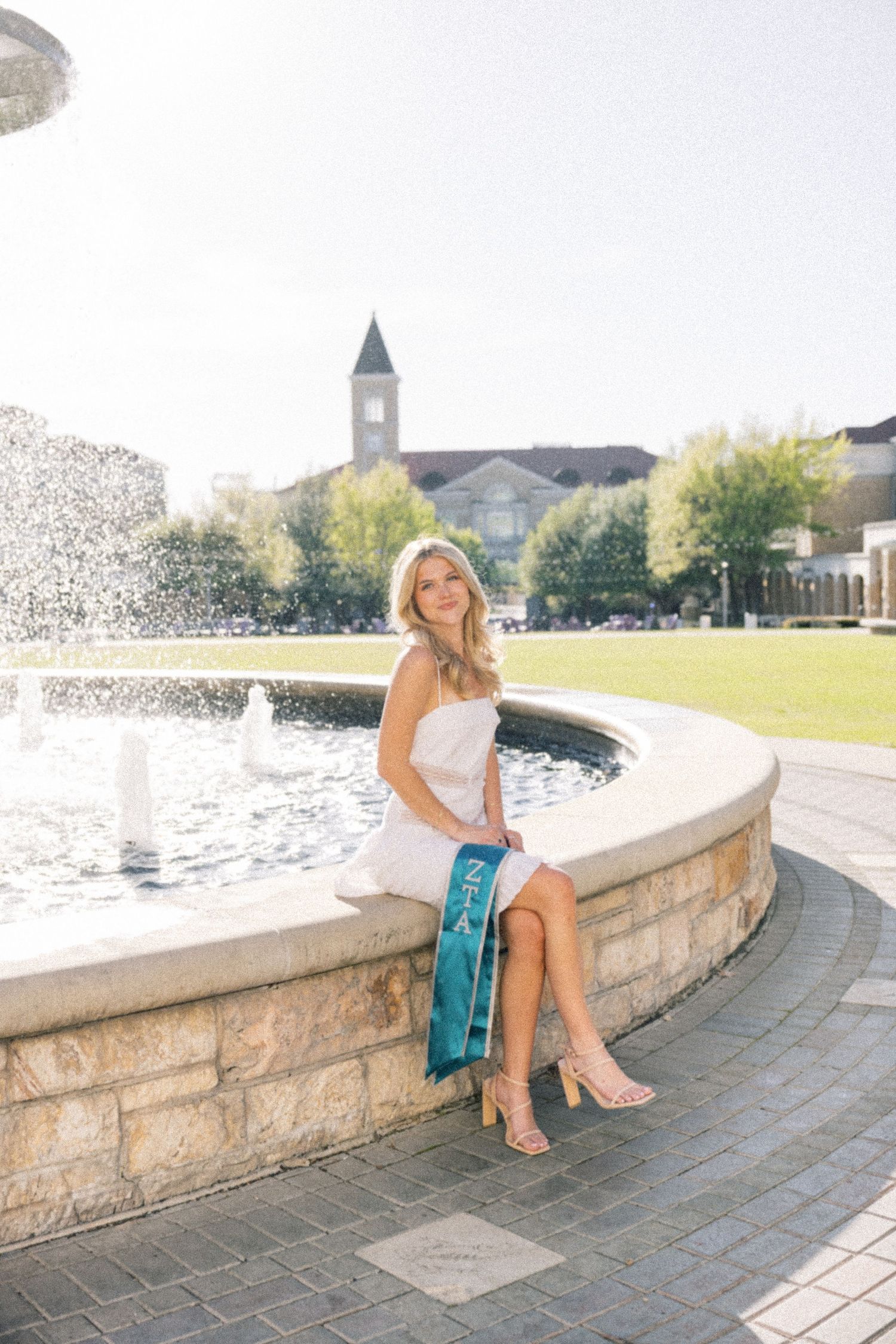 Graduate in white dress with teal sash sitting on fountain edge with campus building behind.
