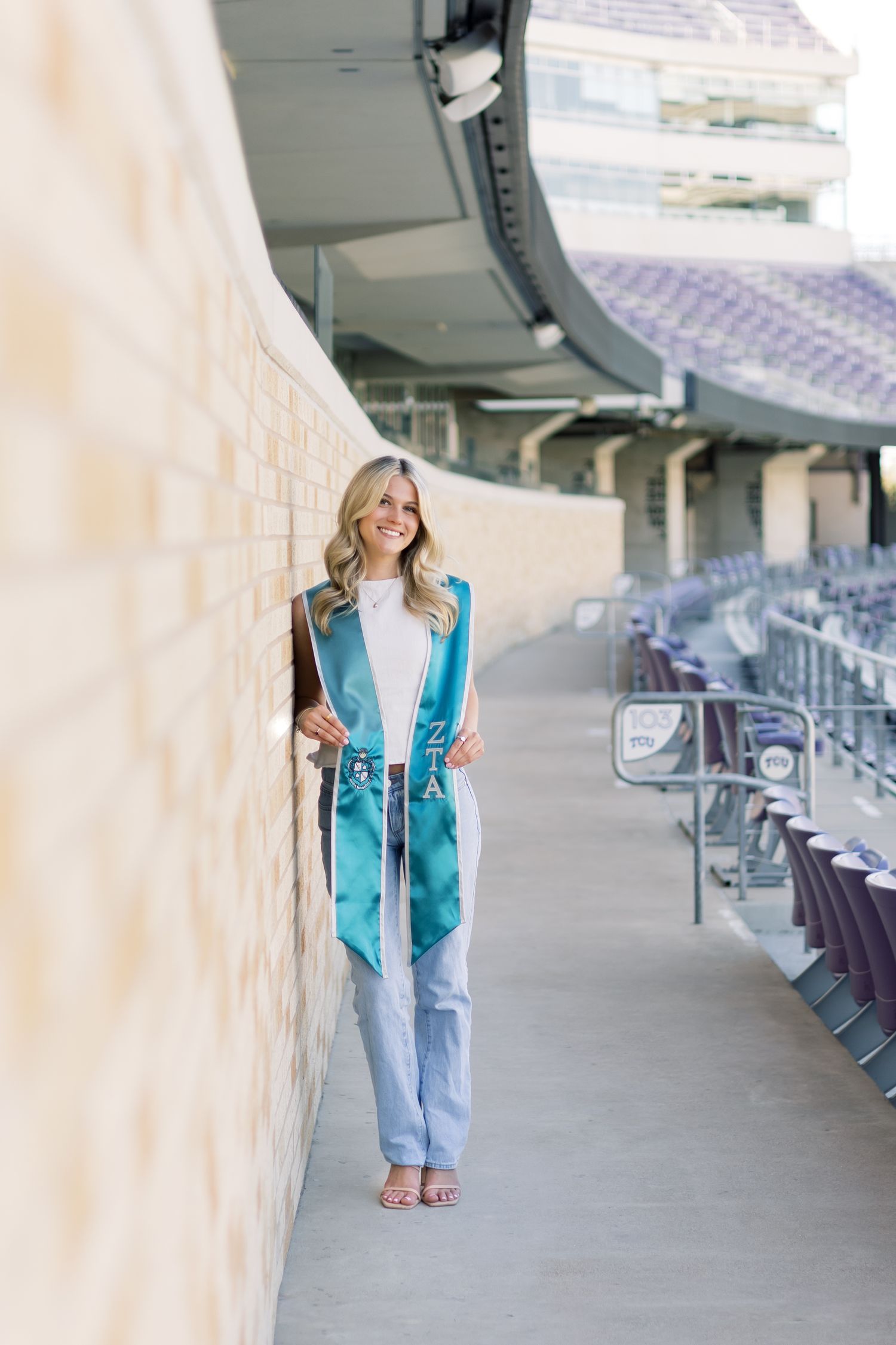 Blonde graduate wearing teal ZTA graduation stole leaning against brick wall at stadium.