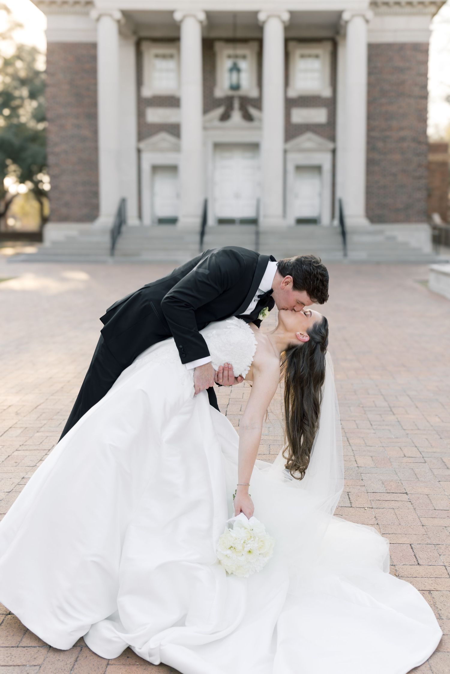 Groom dips bride in stunning white ball gown for a romantic kiss outside a brick colonial building.