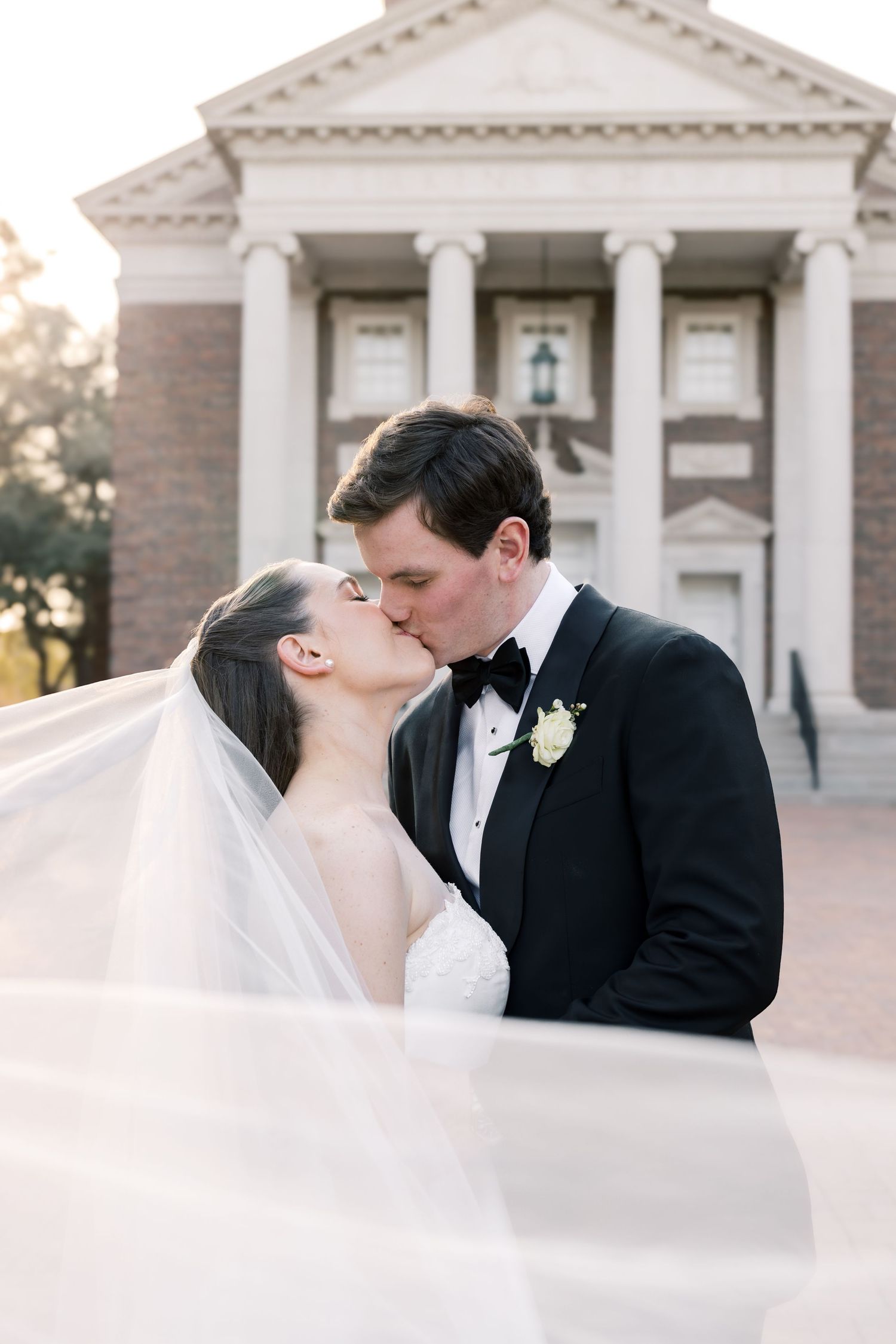 Bride and groom share a tender kiss as her veil billows in the breeze before a brick chapel.