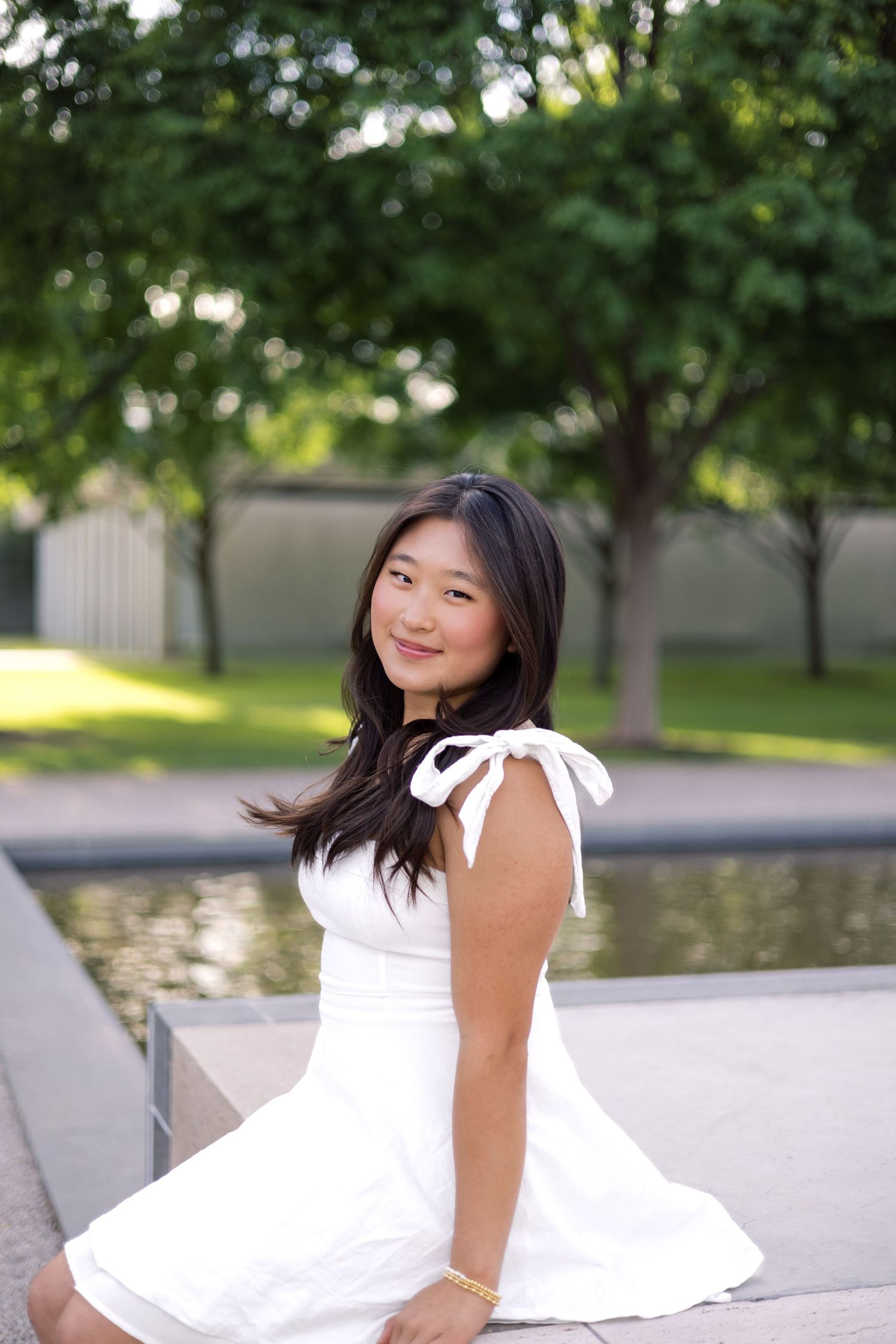 Smiling young woman in white sundress sitting outdoors near a reflecting pool surrounded by trees.
