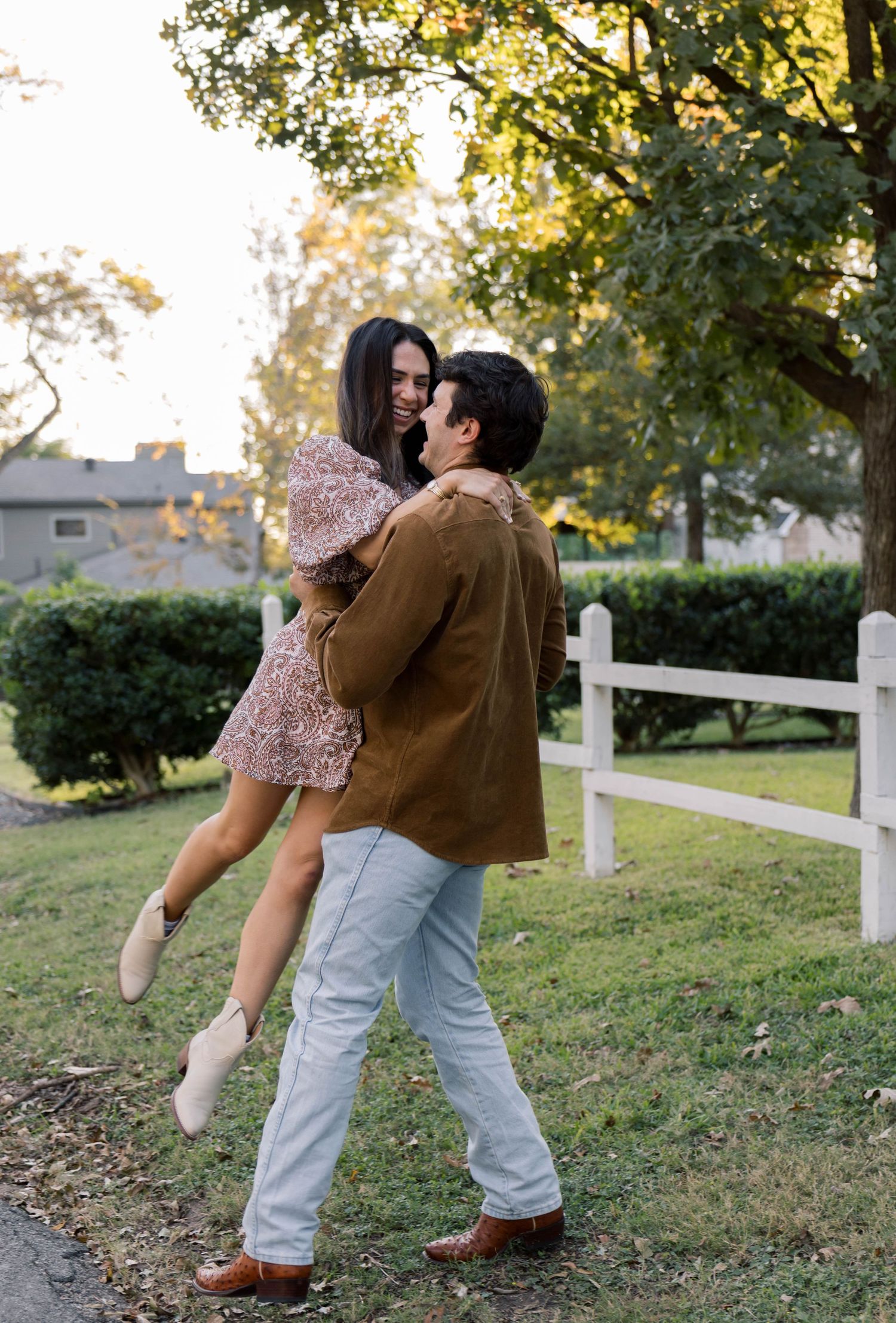 Man lifting woman in floral dress and white boots outdoors near a white fence during golden hour.
