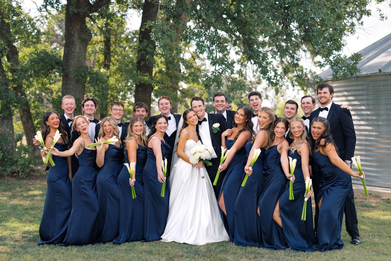 Wedding party in navy blue attire poses outdoors, bride in white gown at center with groom in black tuxedo.