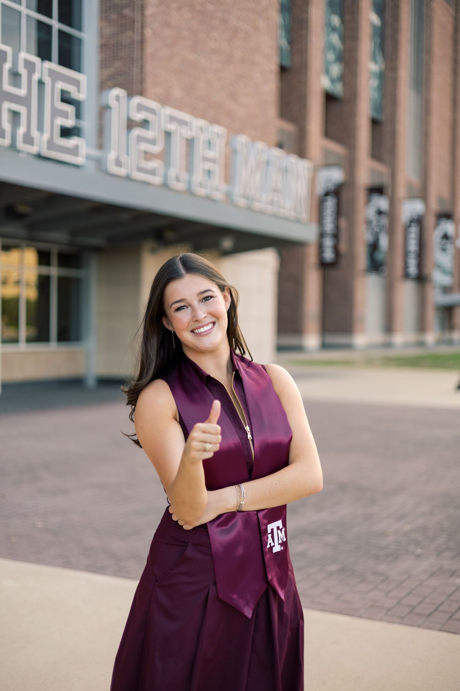 Cheerful Texas A&M graduate in maroon dress giving thumbs up in front of The 12th Man stadium.