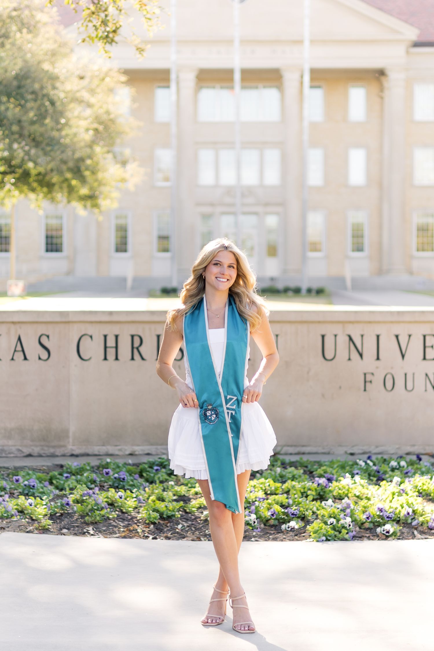 Smiling blonde graduate in white dress with ZTA teal stole posing at Texas Christian University sign.