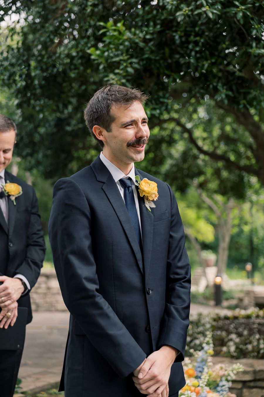 Groom in navy suit with yellow rose boutonniere smiling joyfully outdoors at wedding ceremony.