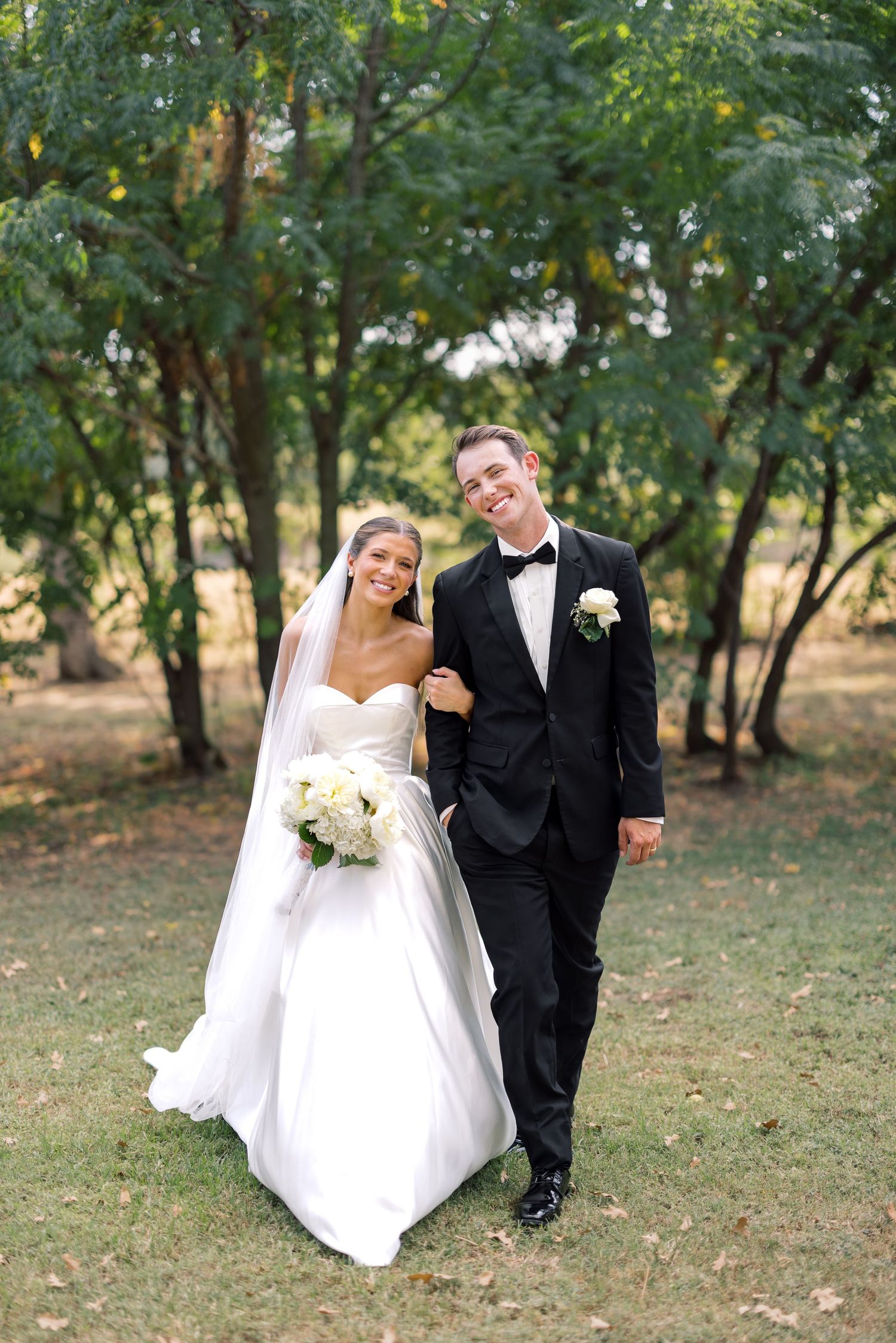 Smiling bride in white gown and groom in black tuxedo walk together outdoors among green trees on their wedding day.