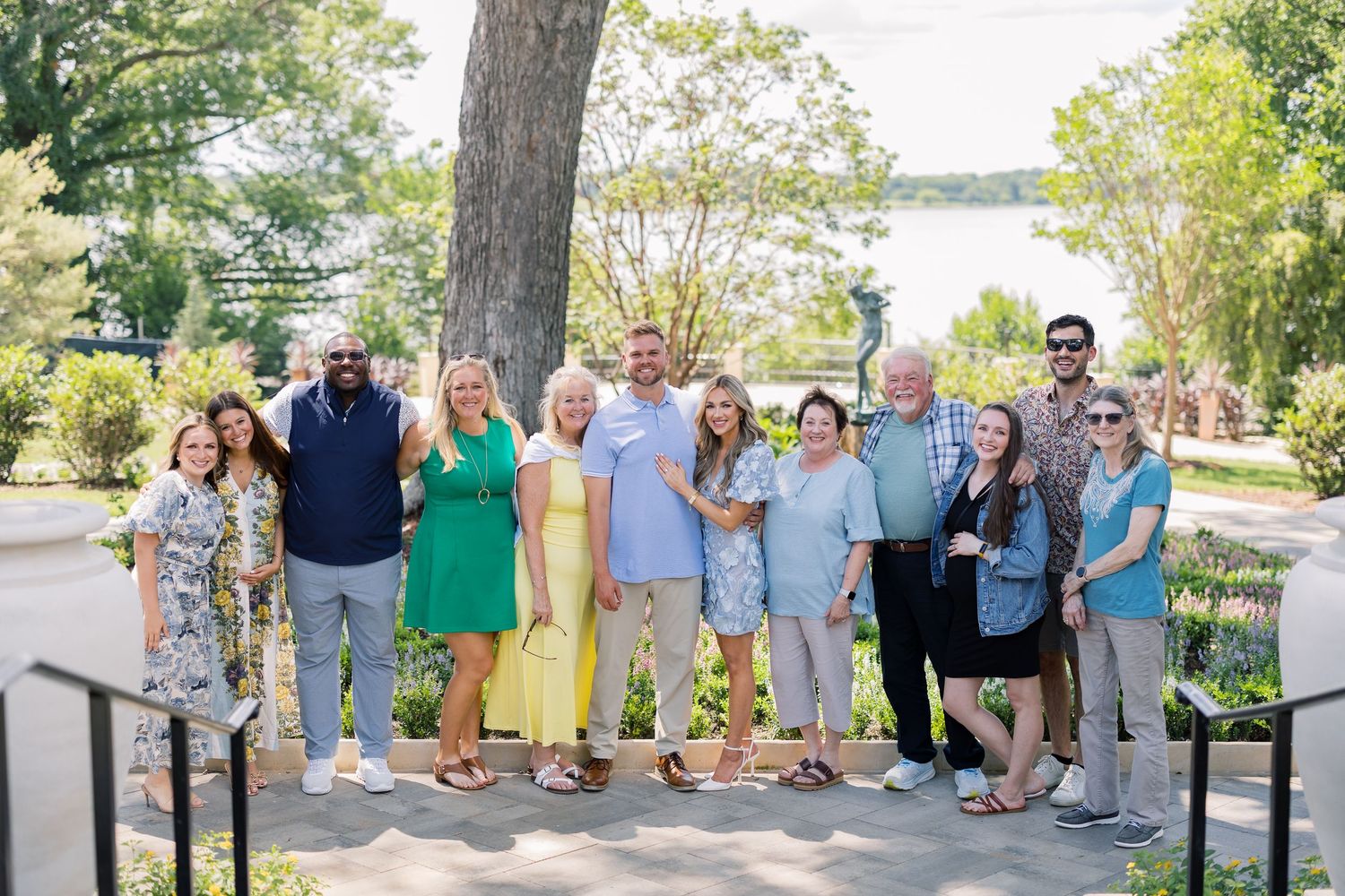 Large group of smiling adults posing outdoors near a tree with a lake and greenery in the background.