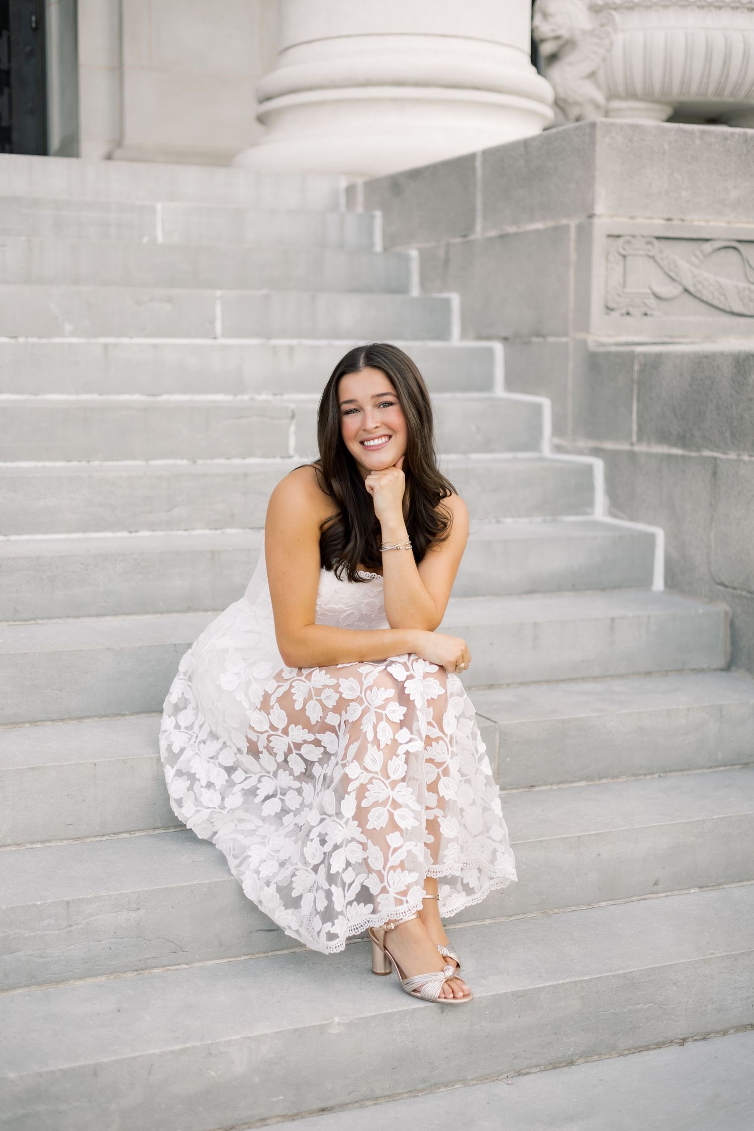 Smiling woman in white lace midi dress seated on marble steps with columns behind her.