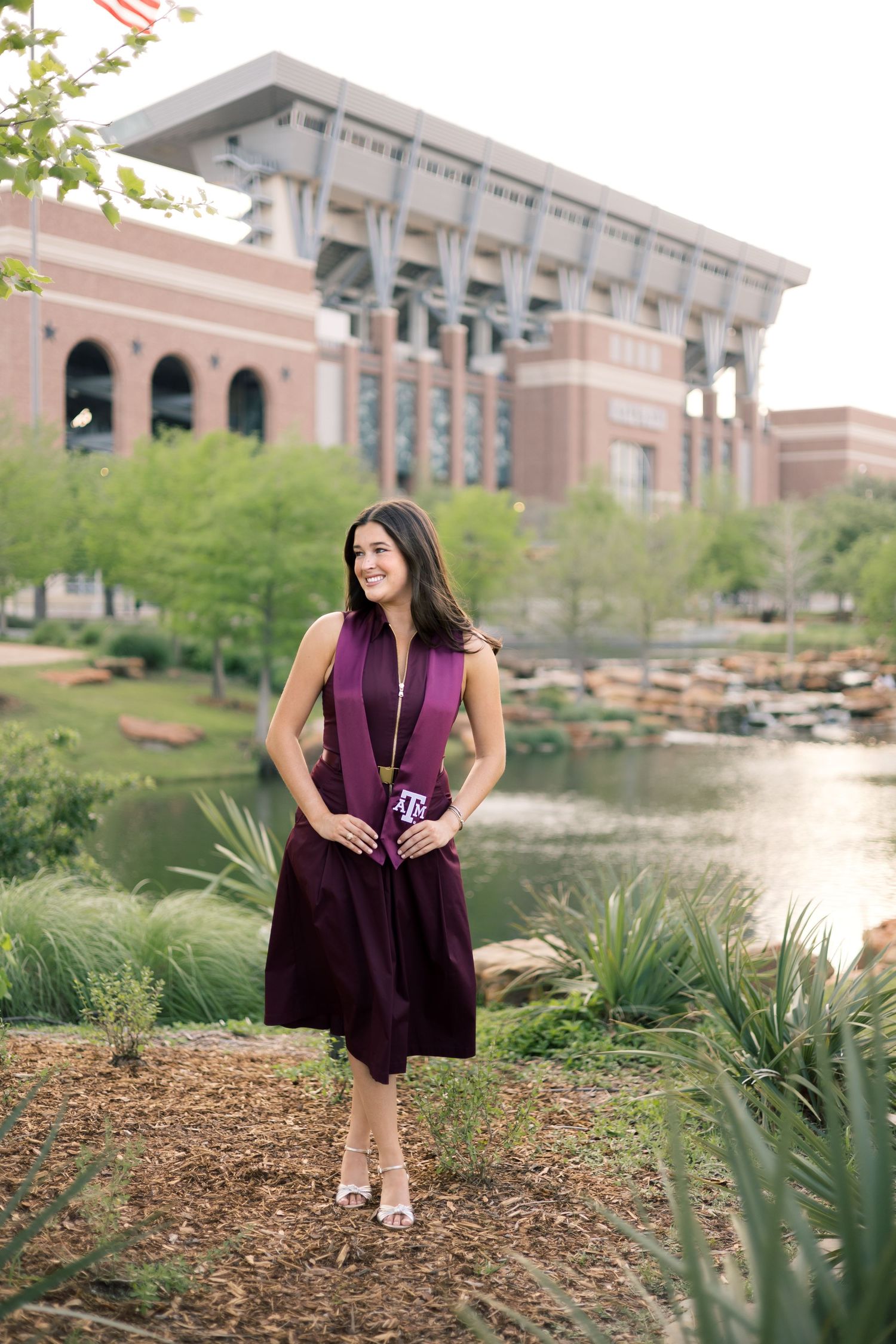 Texas A&M graduate in maroon dress stands by pond with stadium in background.