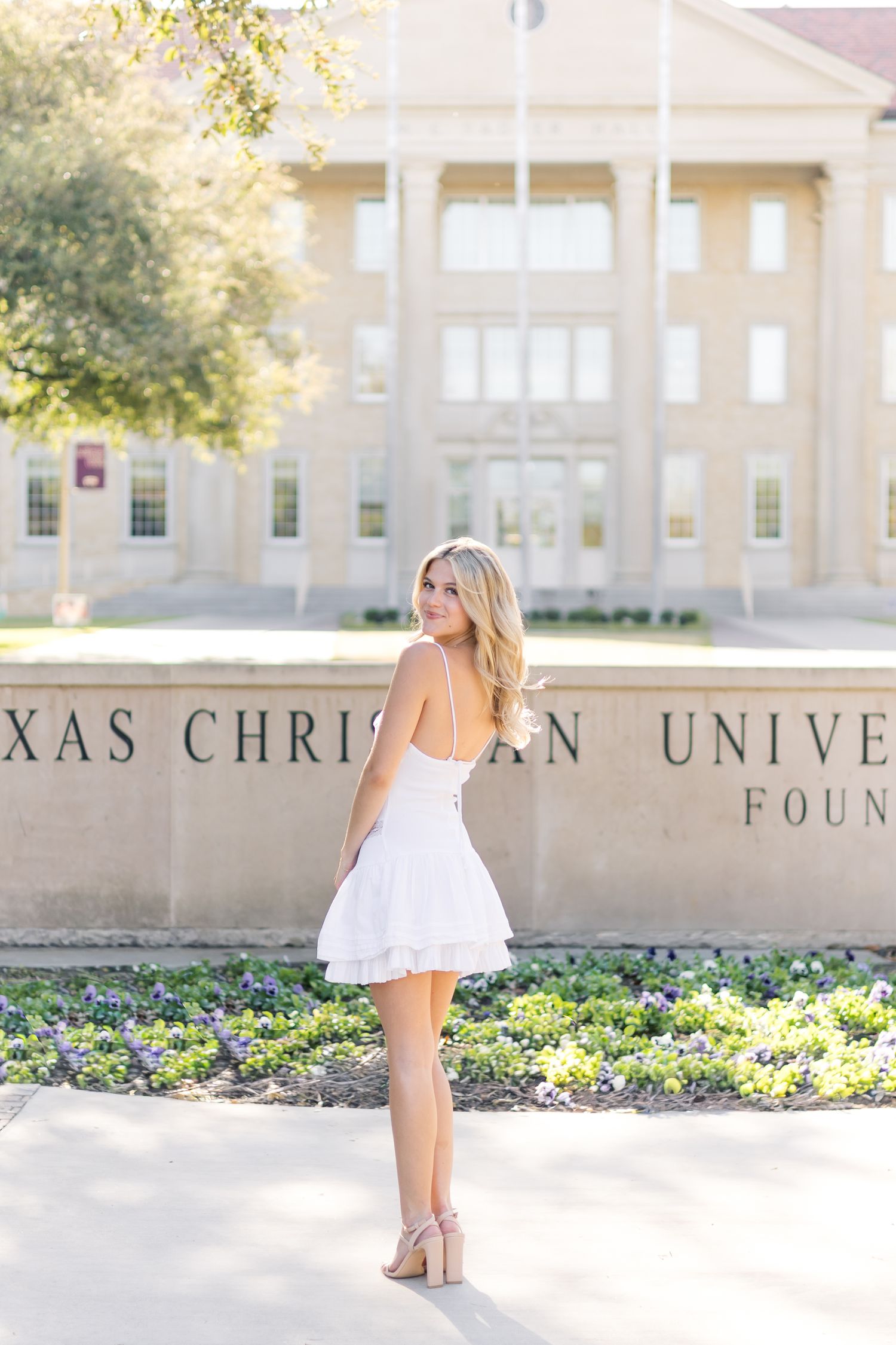 Blonde woman in white dress posing in front of Texas Christian University Founded sign.