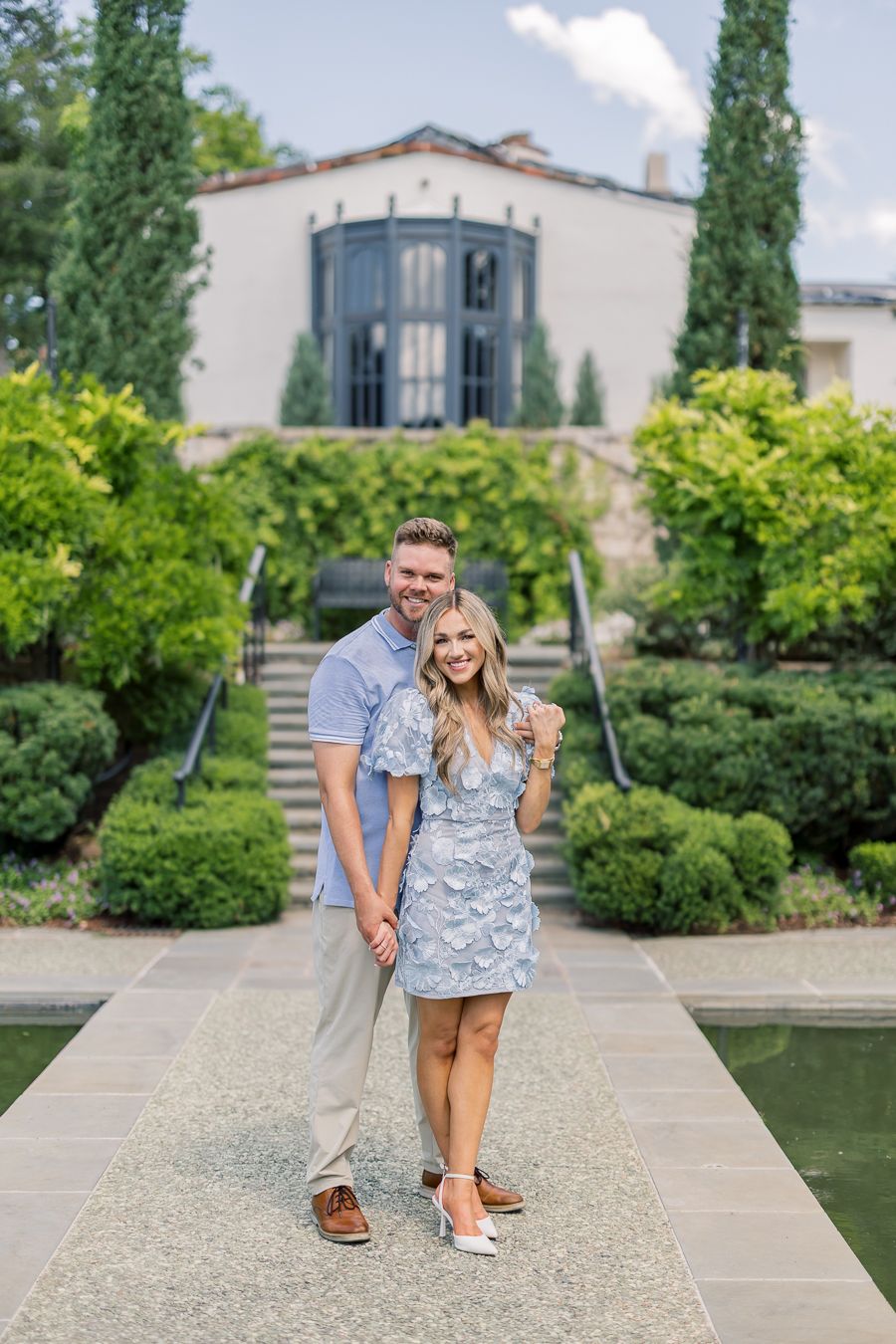 Smiling couple poses in front of elegant garden fountain, woman in blue floral dress shows engagement ring.