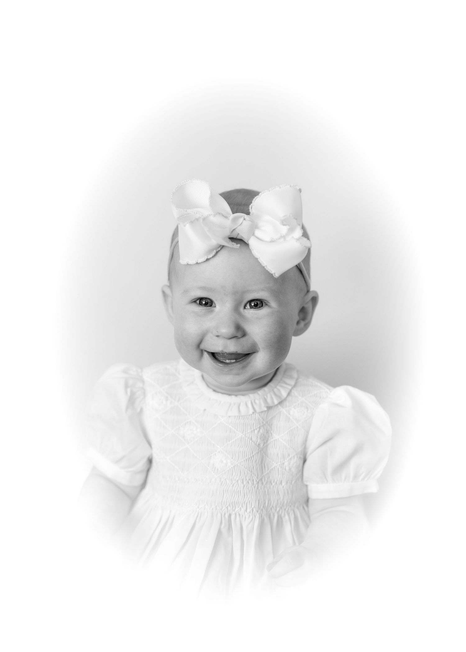 Black and white portrait of a smiling baby girl wearing a white dress and large bow headband.