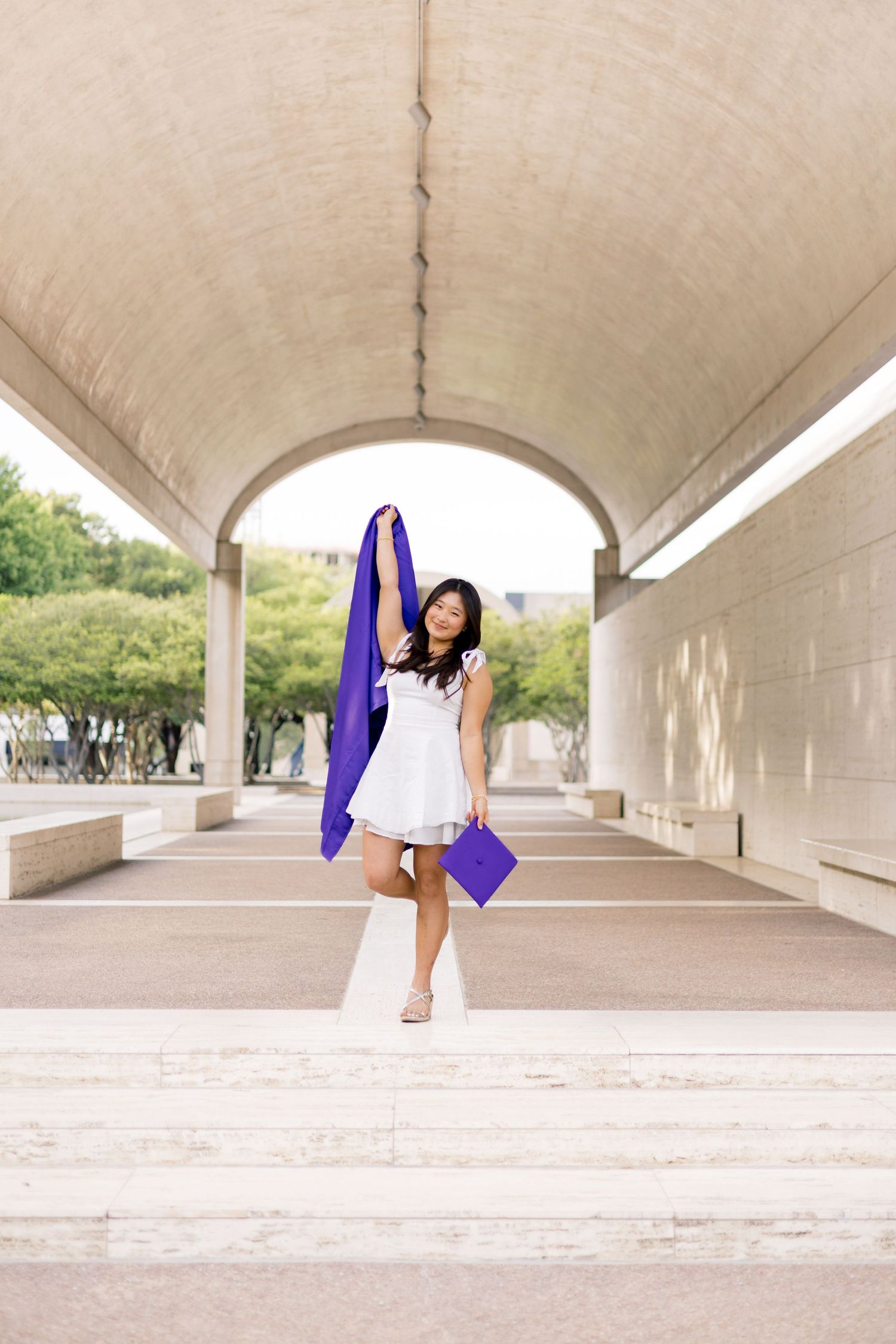 Young woman in white dress celebrating graduation, holding purple cap and gown under an arched walkway outdoors.