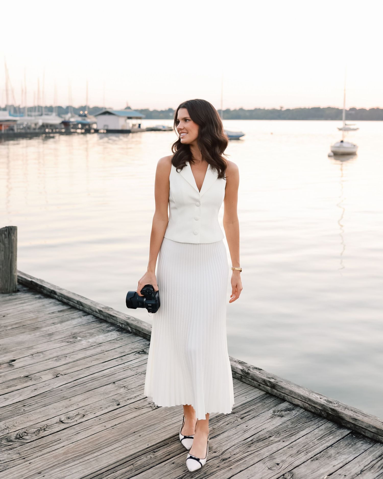 Female photographer in white vest and pleated skirt holding camera on wooden dock at sunset by marina.