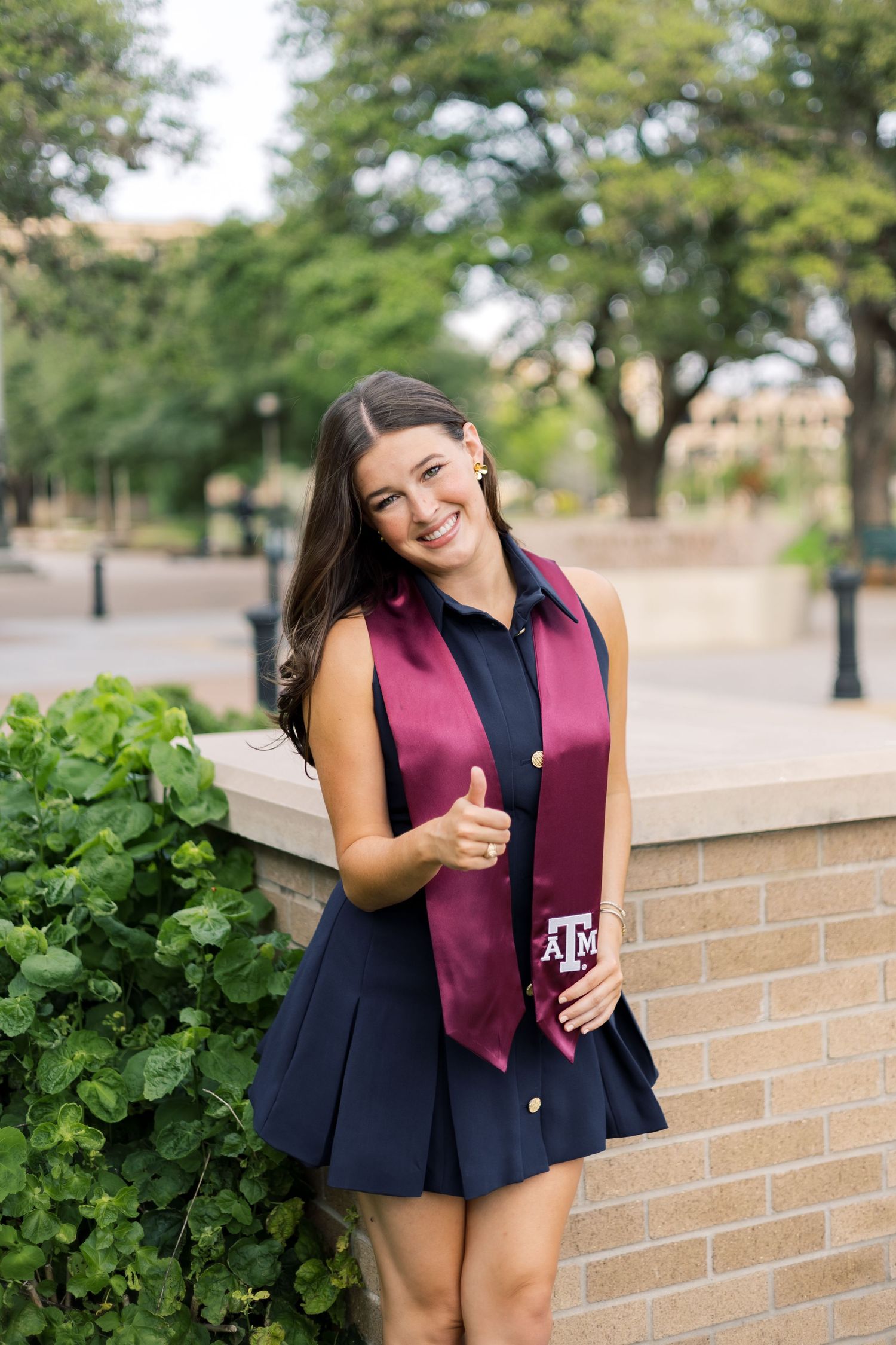 Cheerful Texas A&M graduate gives thumbs up in navy dress with maroon ATM stole near brick wall.