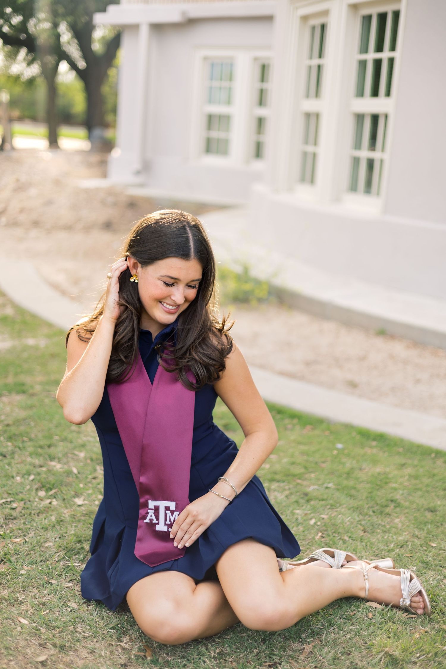 Texas A&M senior in navy dress and maroon stole sits on grass adjusting hair near campus building.