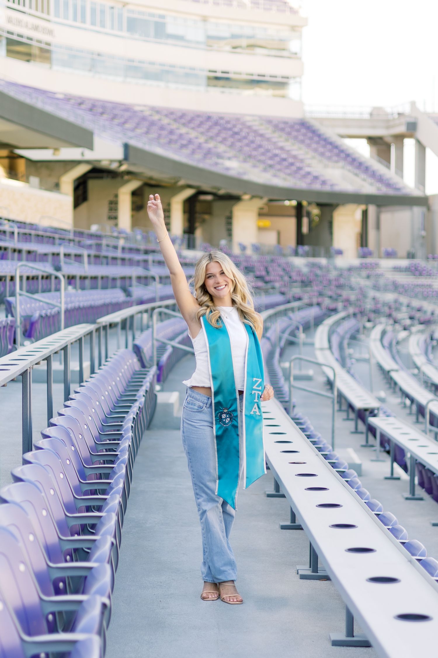 Smiling blonde woman raising arm wearing ZTA teal graduation stole in purple TCU stadium stands.