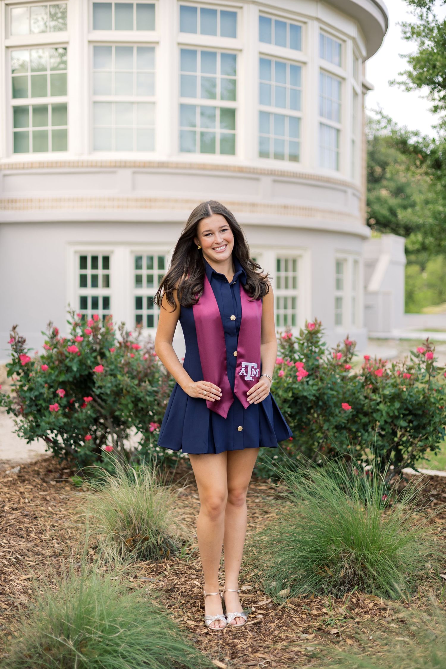 Brunette woman in navy blue dress wearing Texas A&M maroon graduation stole standing near rose bushes outdoors.
