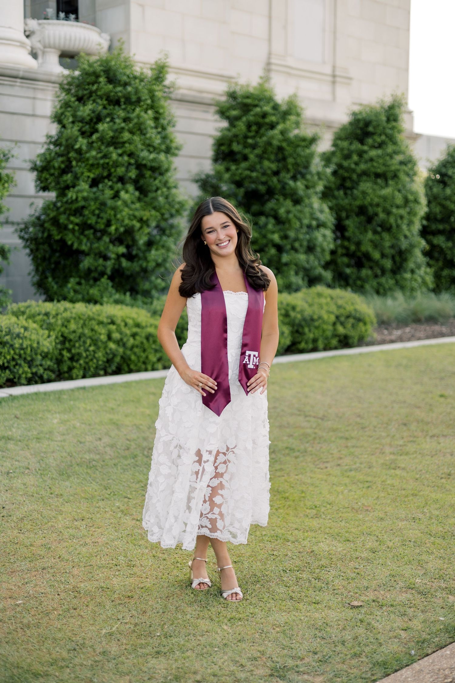 Graduate in white lace dress wearing maroon Texas A&M stole standing on green lawn outside building.